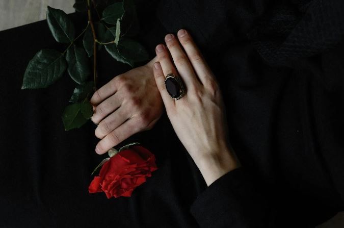 Hands holding a red rose and wearing a dark ring on a black background.
