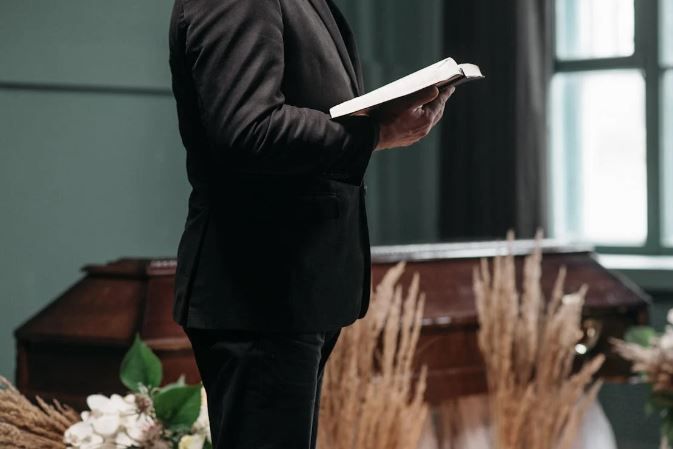 Person in black suit reading from a book at a funeral, with a casket and floral arrangements.