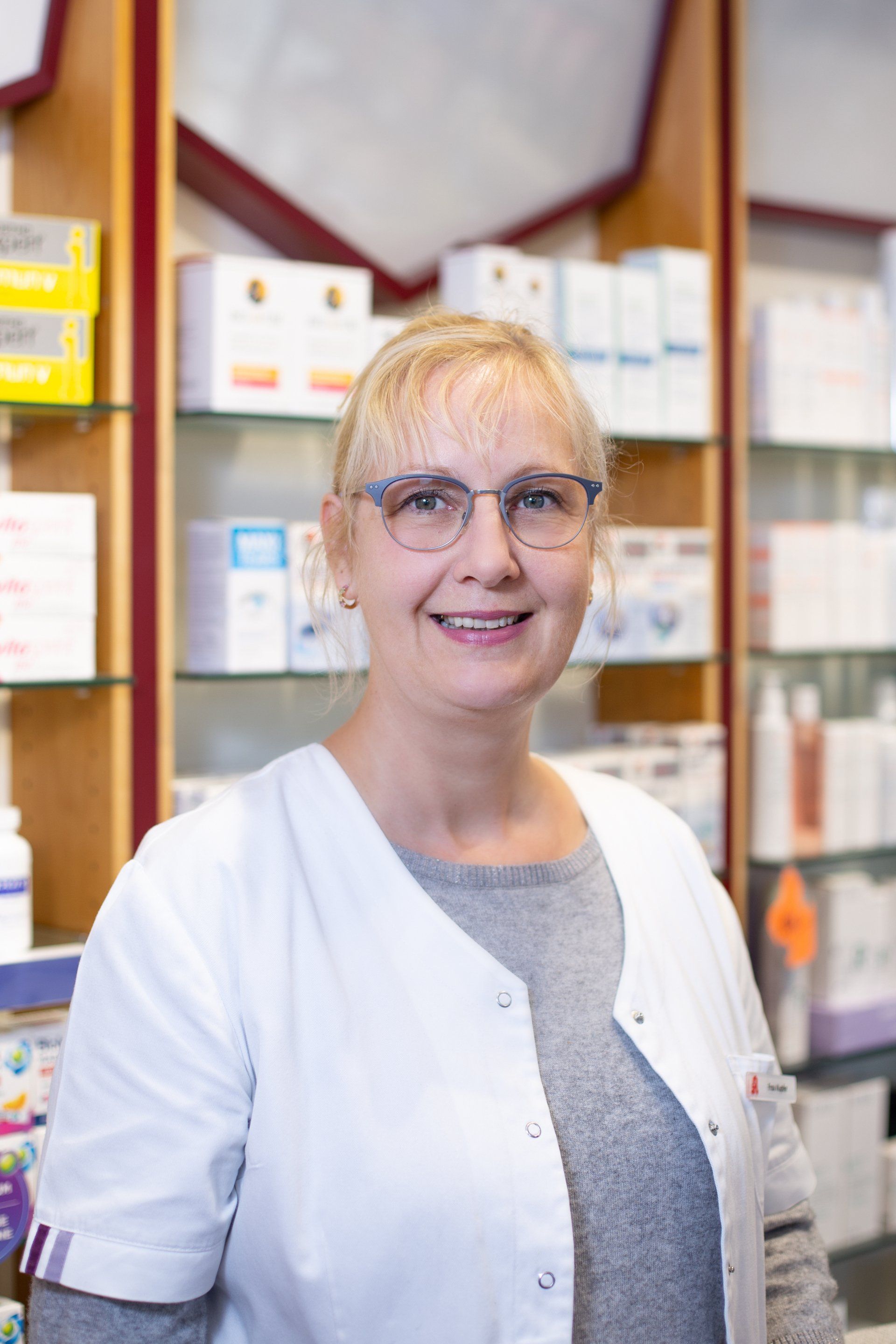Eine Frau in einem weißen Kittel und mit Brille steht in einer Apotheke.