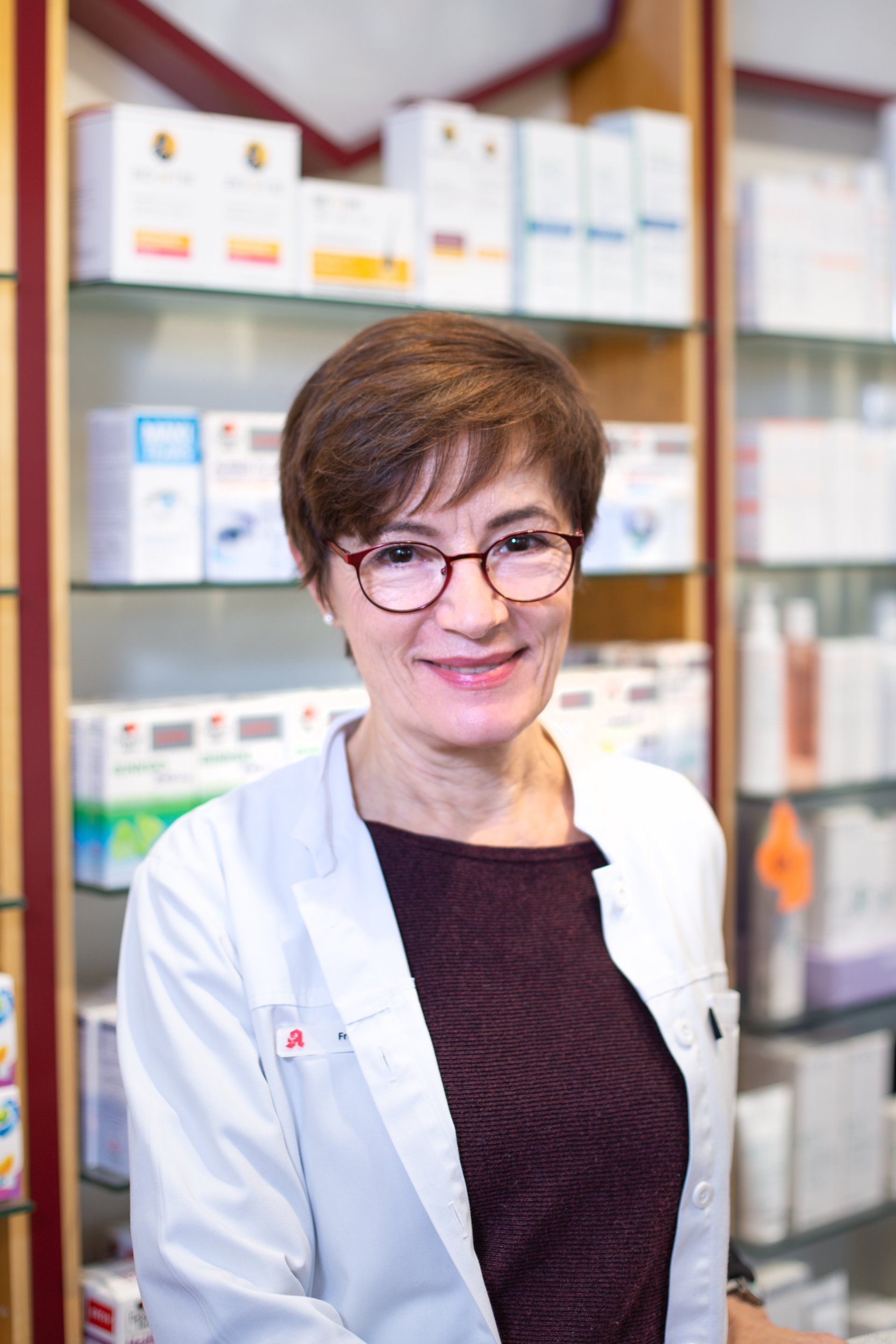 Eine Frau mit Brille und weißem Kittel steht vor einem Regal in einer Apotheke.