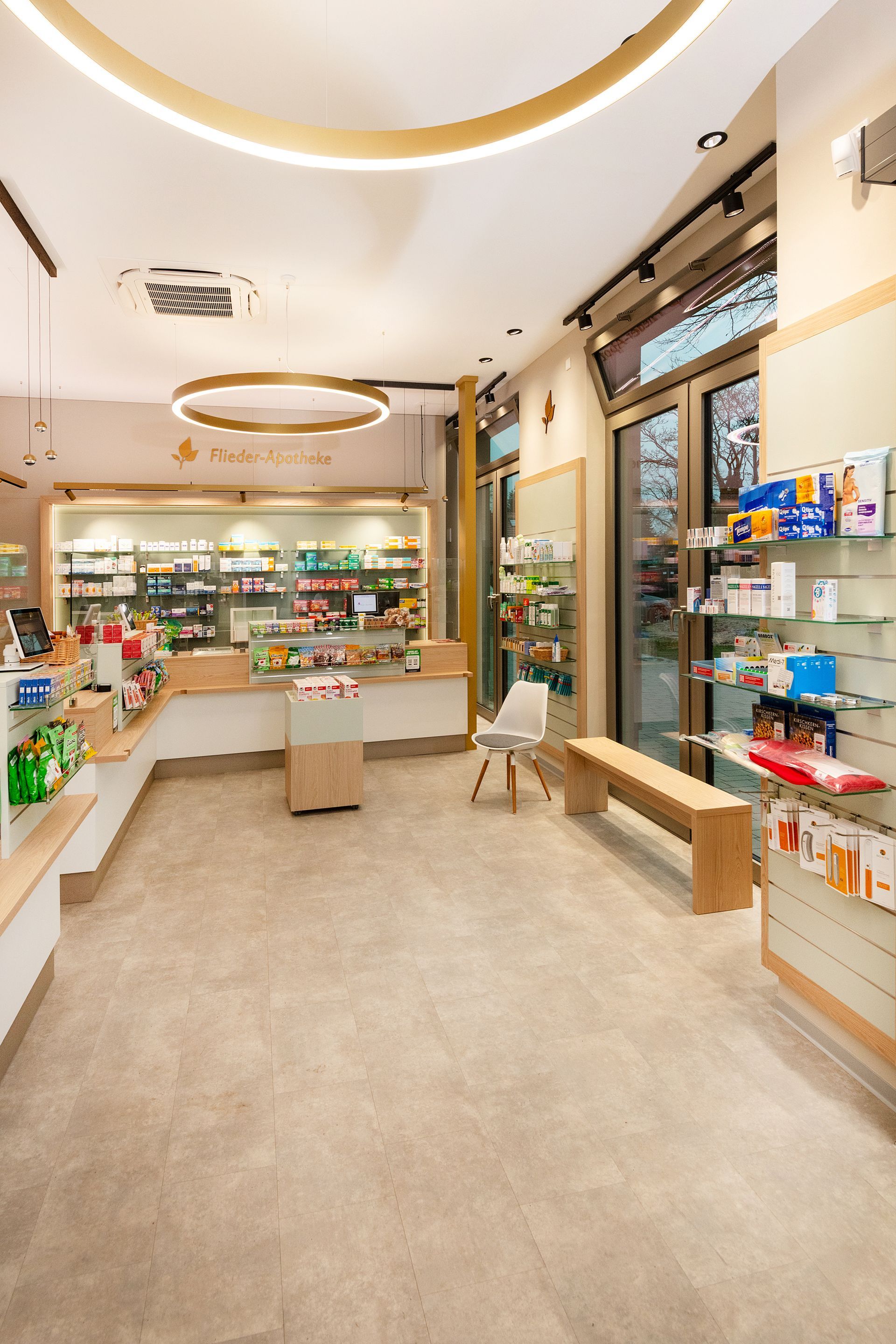 Innenansicht einer Apotheke mit Regalen voller Produkte, einem Tresen und Sitzgelegenheiten. Gut beleuchtet in neutralen Farbtönen.