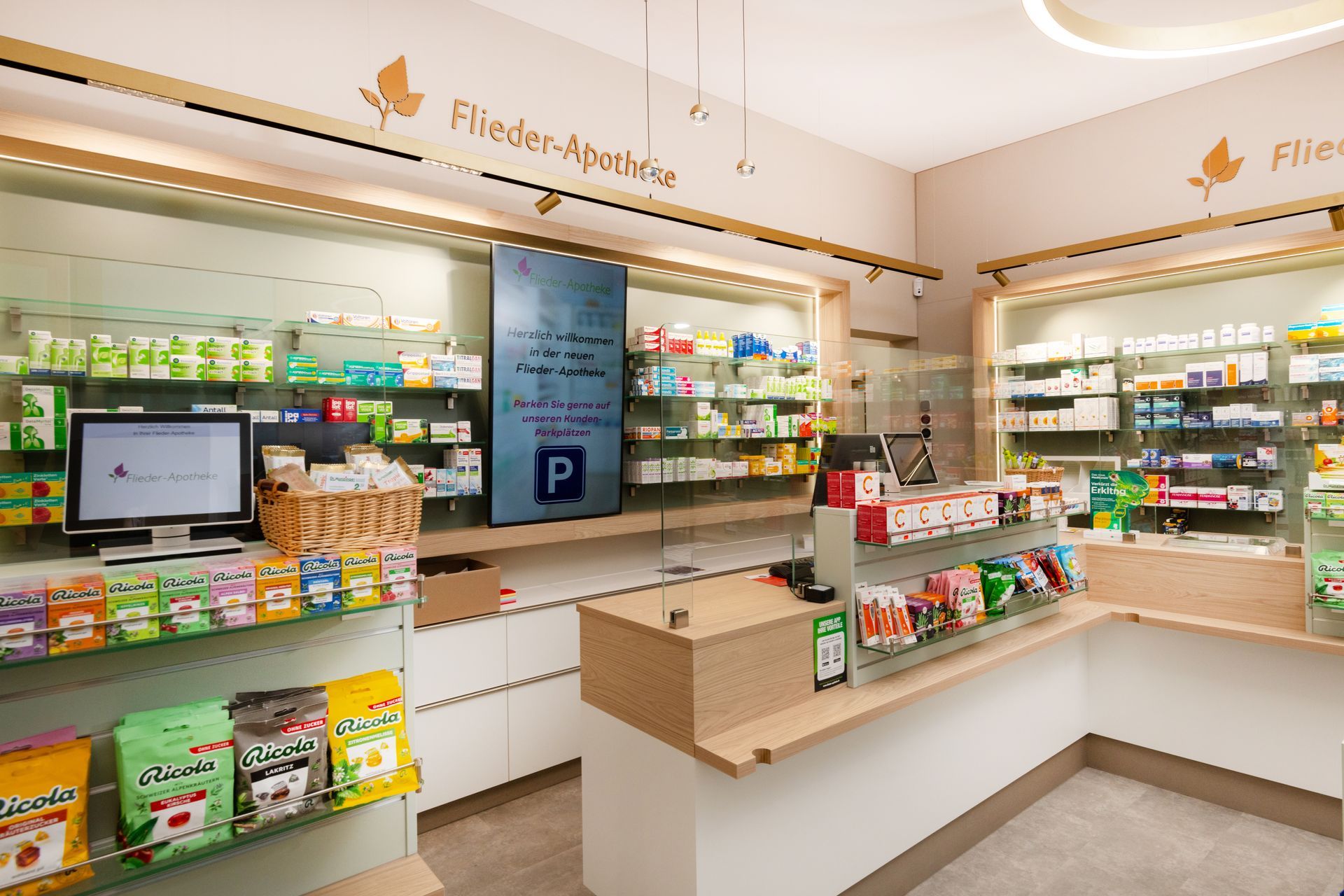 Innenansicht einer Apotheke mit Regalen voller Produkte, einem Tresen und einem digitalen Display. Helle Holz- und beigefarbene Dekoration.