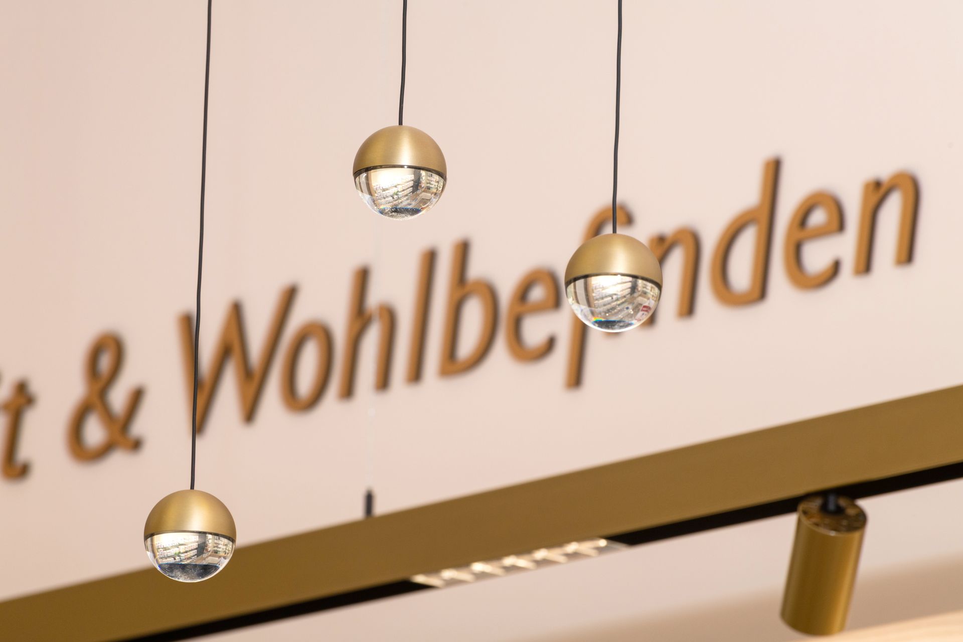 Drei Pendelleuchten mit goldenen Oberteilen und gemusterten Glaskugeln vor einer verschwommenen Wand mit Text.