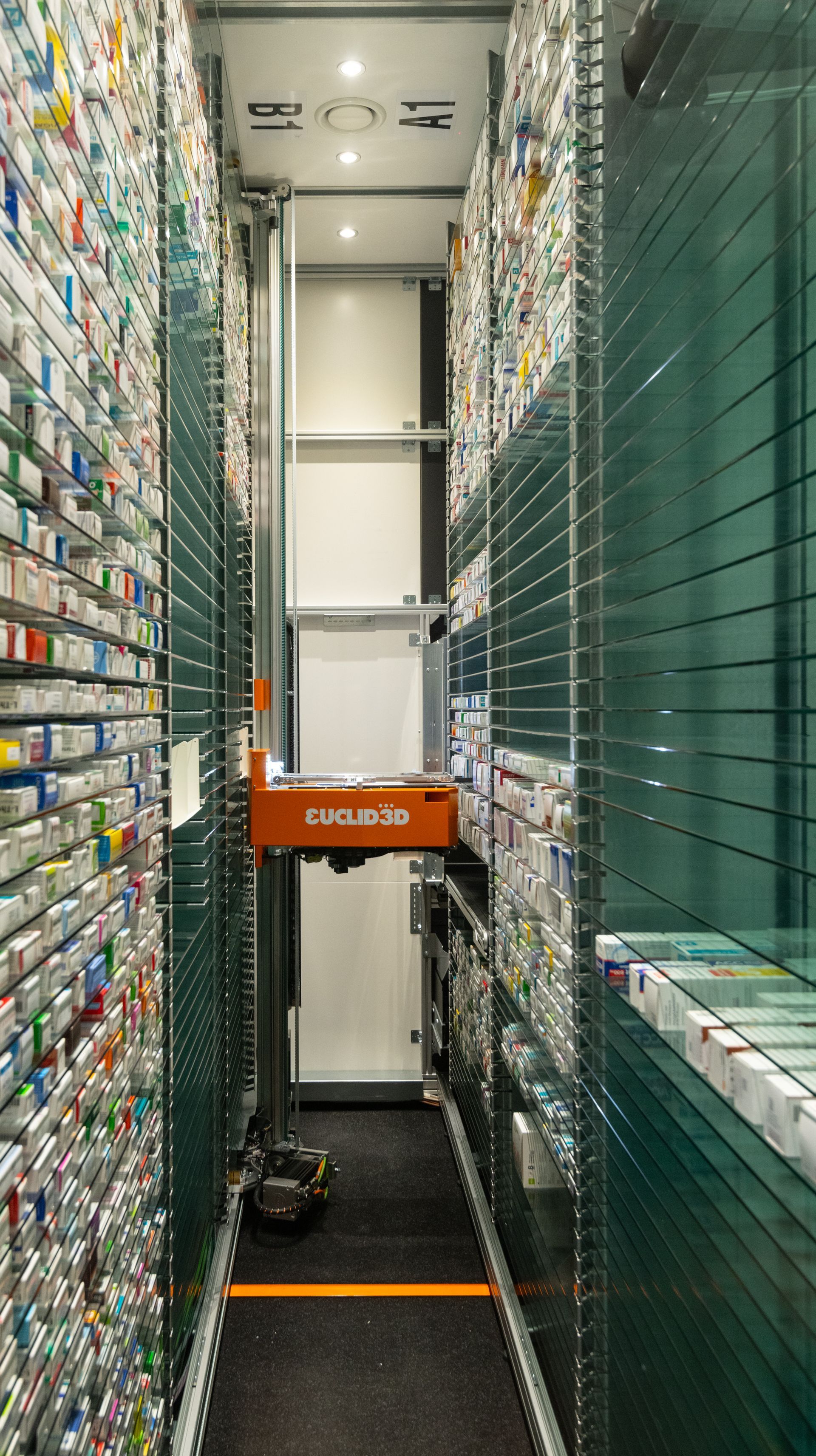 Automatisiertes Apothekenregalsystem mit orangefarbenem Roboter zur Medikamentenentnahme in einem schmalen Gang.