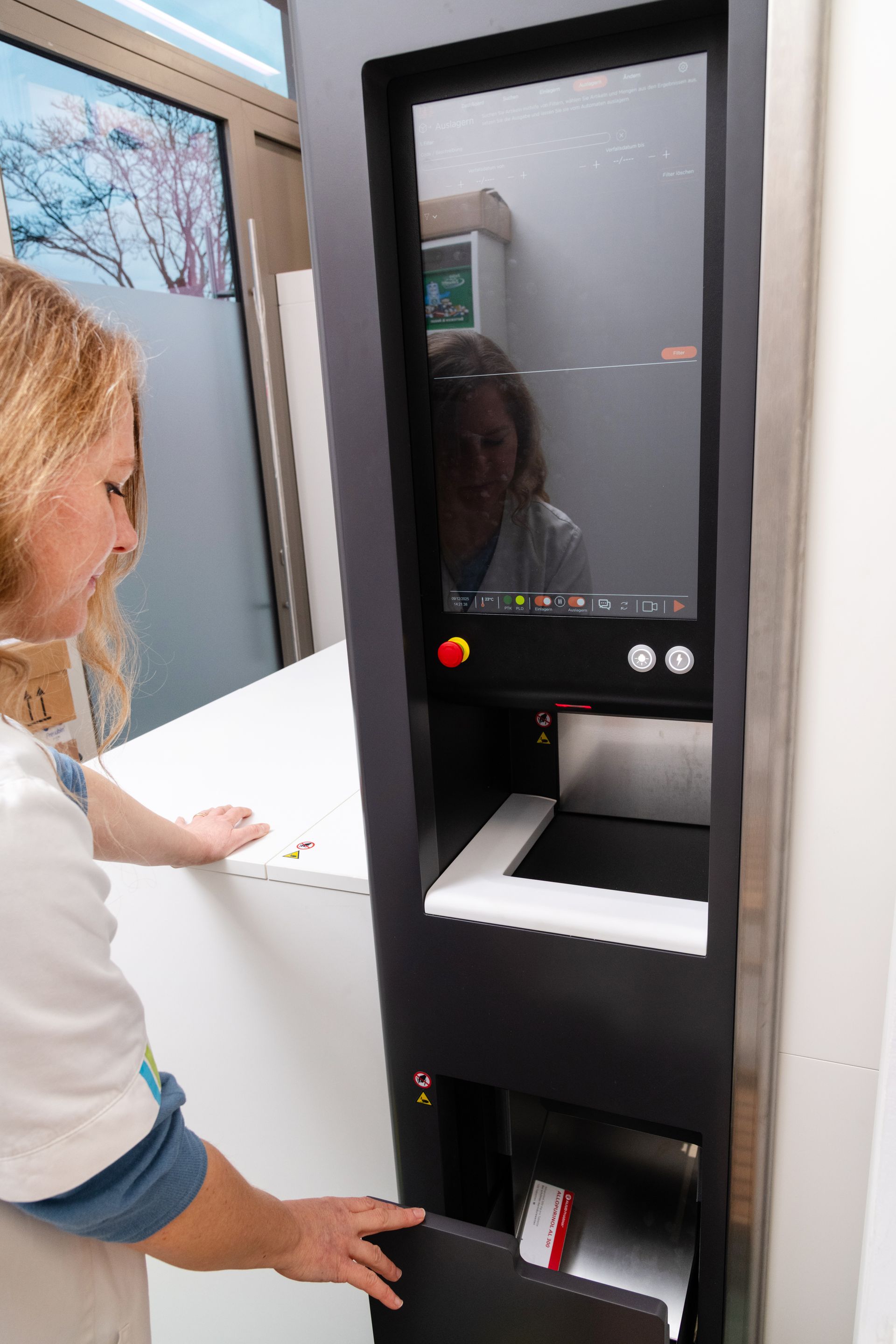 Eine Frau öffnet die untere Tür eines schwarzen Medizinschranks mit einem großen Bildschirm; im Bildschirm spiegelt sich eine zweite Person.