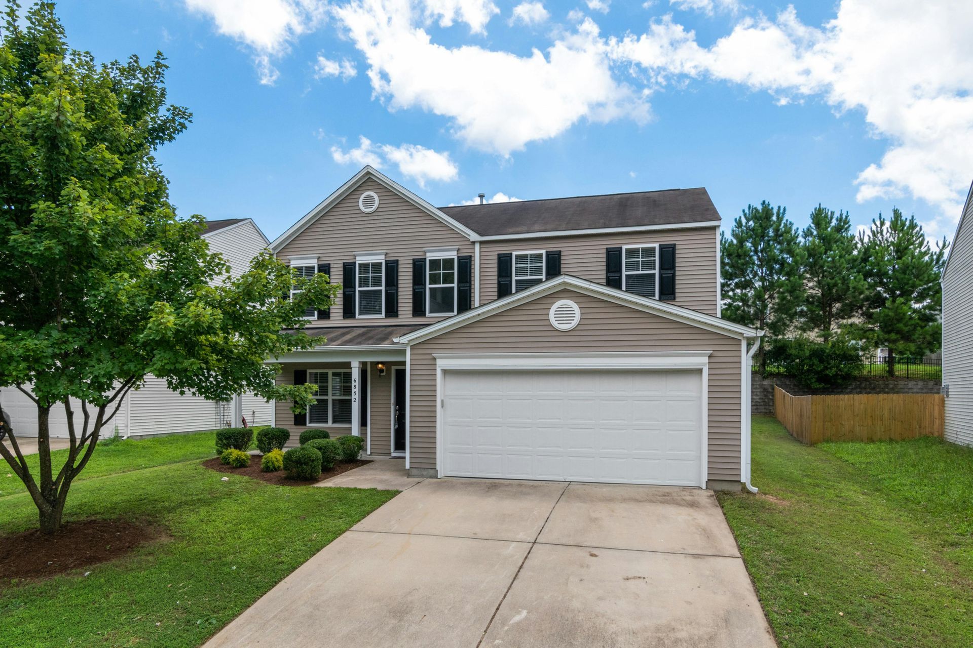 Two-story beige house with a two-car garage, black shutters, and green lawn under a blue, cloudy sky.