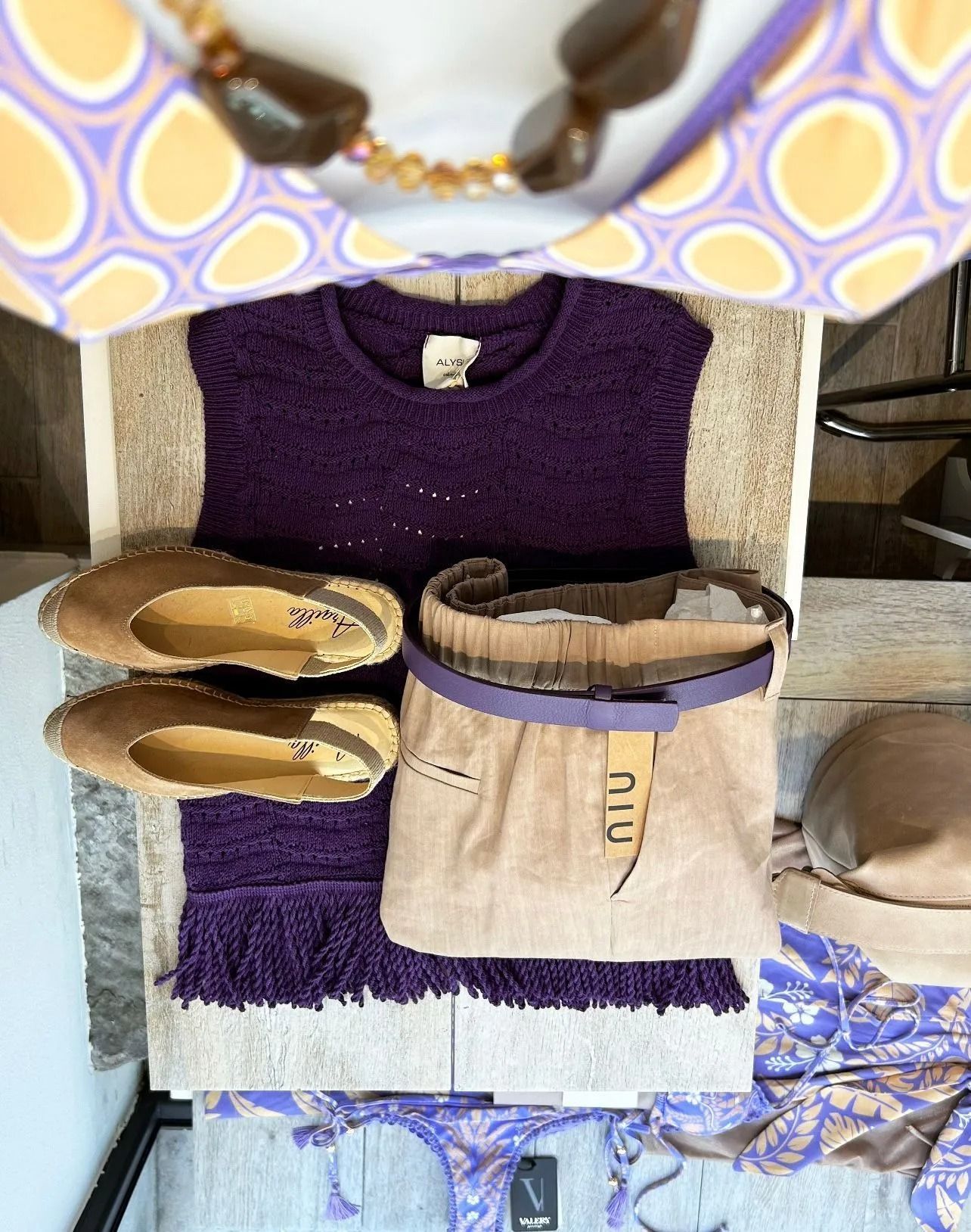 Top viola lavorato a maglia, pantaloni color cuoio ed espadrillas disposti su un tavolo, con sopra un top fantasia e gioielli.