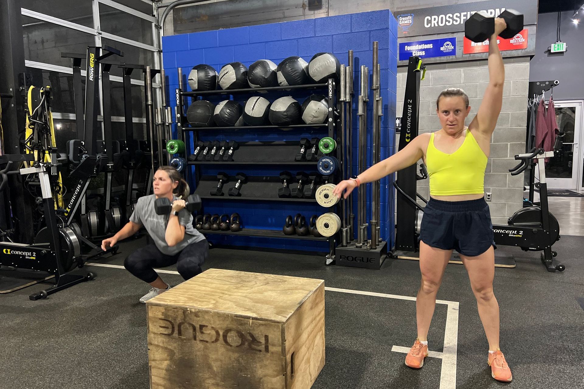 Two women are squatting and lifting dumbbells in a gym.