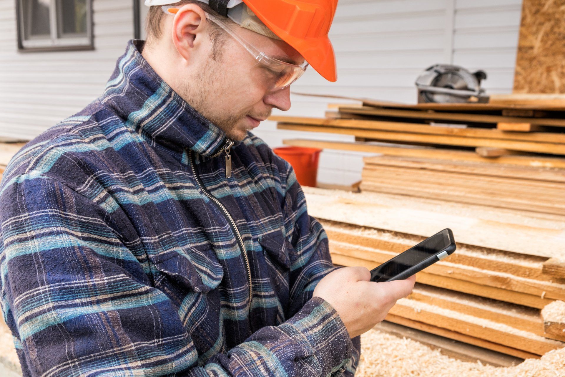Ouvrier du bâtiment portant un casque orange et des lunettes de sécurité utilisant un smartphone, avec du bois en arrière-plan.