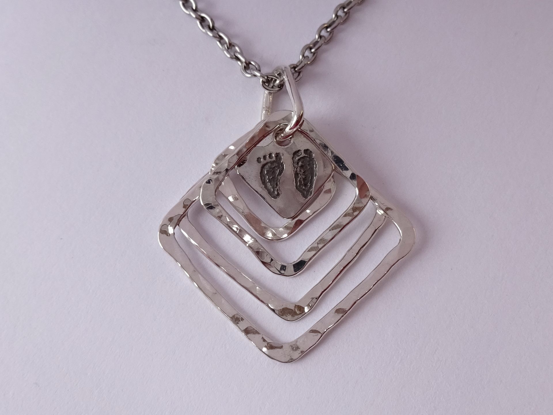Zilveren hanger met gehamerde open vierkanten met voetafdrukken en letters aan de achterkant.