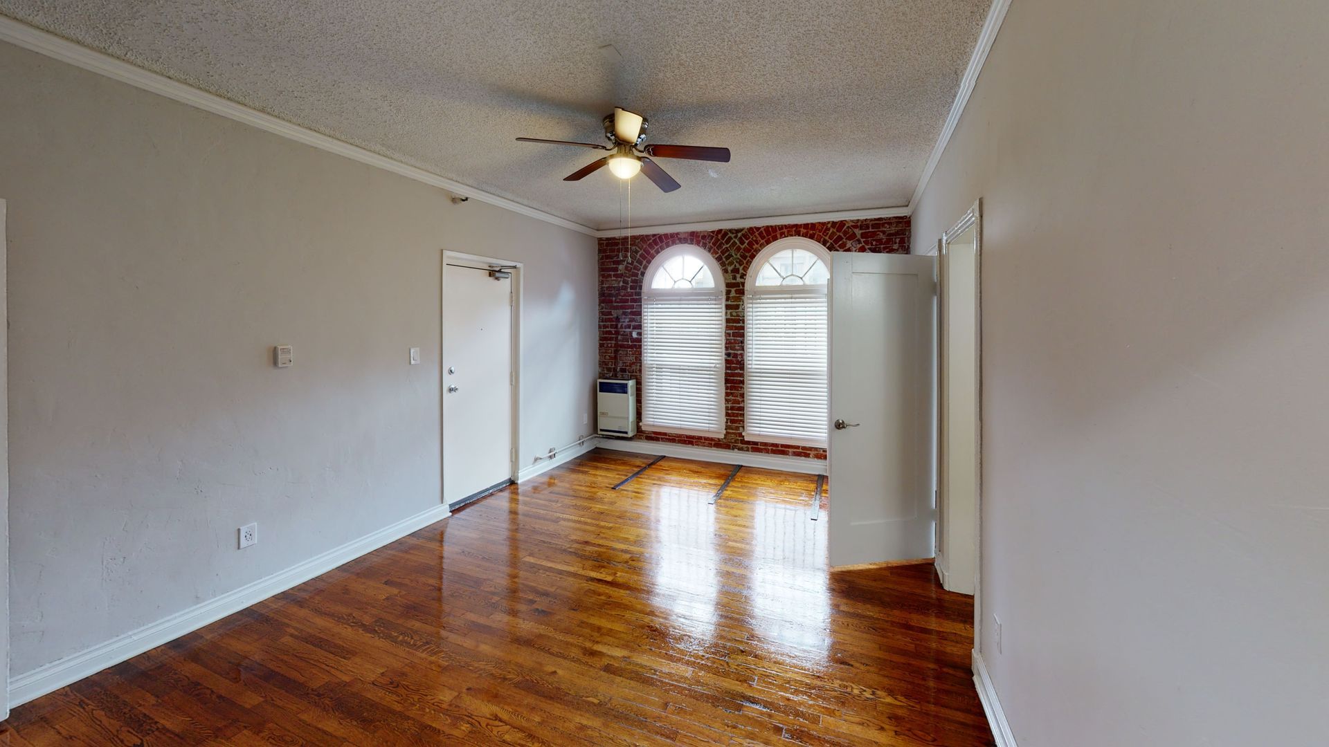 Empty living room with wood floors, brick accent wall, and arched windows.