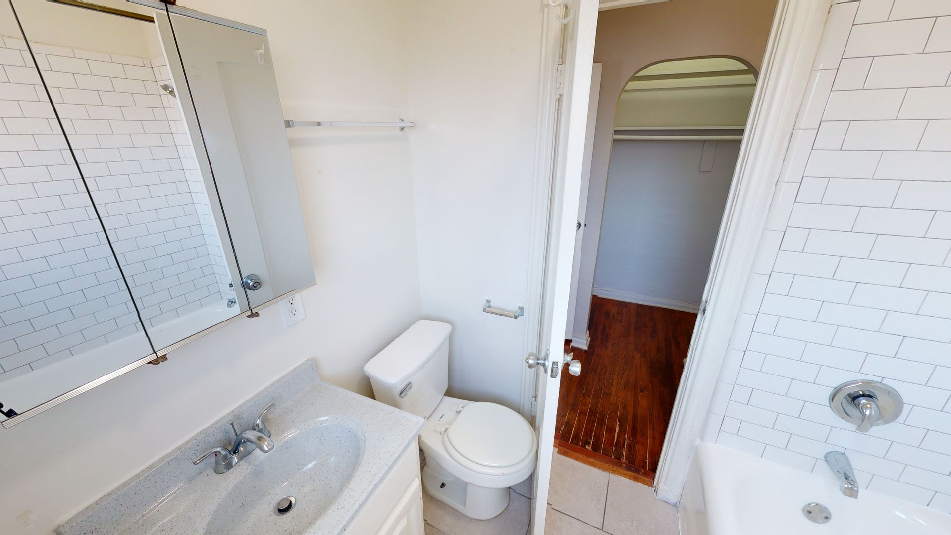 Small, white bathroom with a toilet, sink, mirror, and an open doorway to a dark room with a closet.