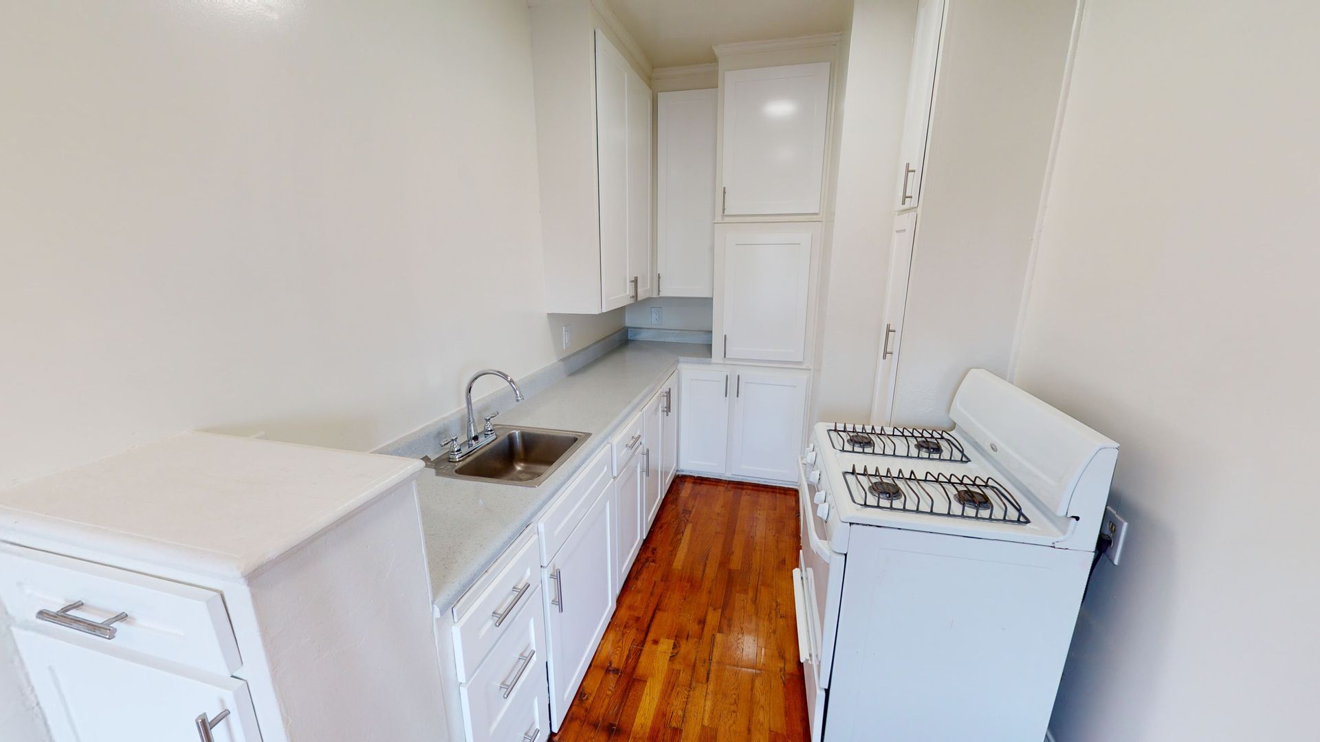 Narrow kitchen with white cabinets, silver sink, and stove with wooden floor.