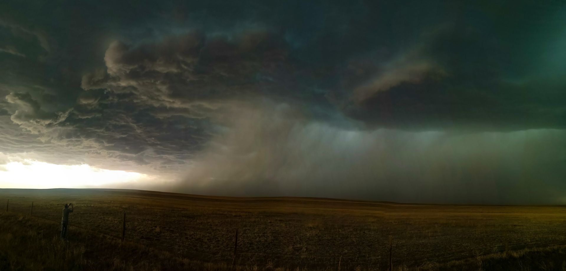 A storm is coming in over a field with a fence in the foreground.