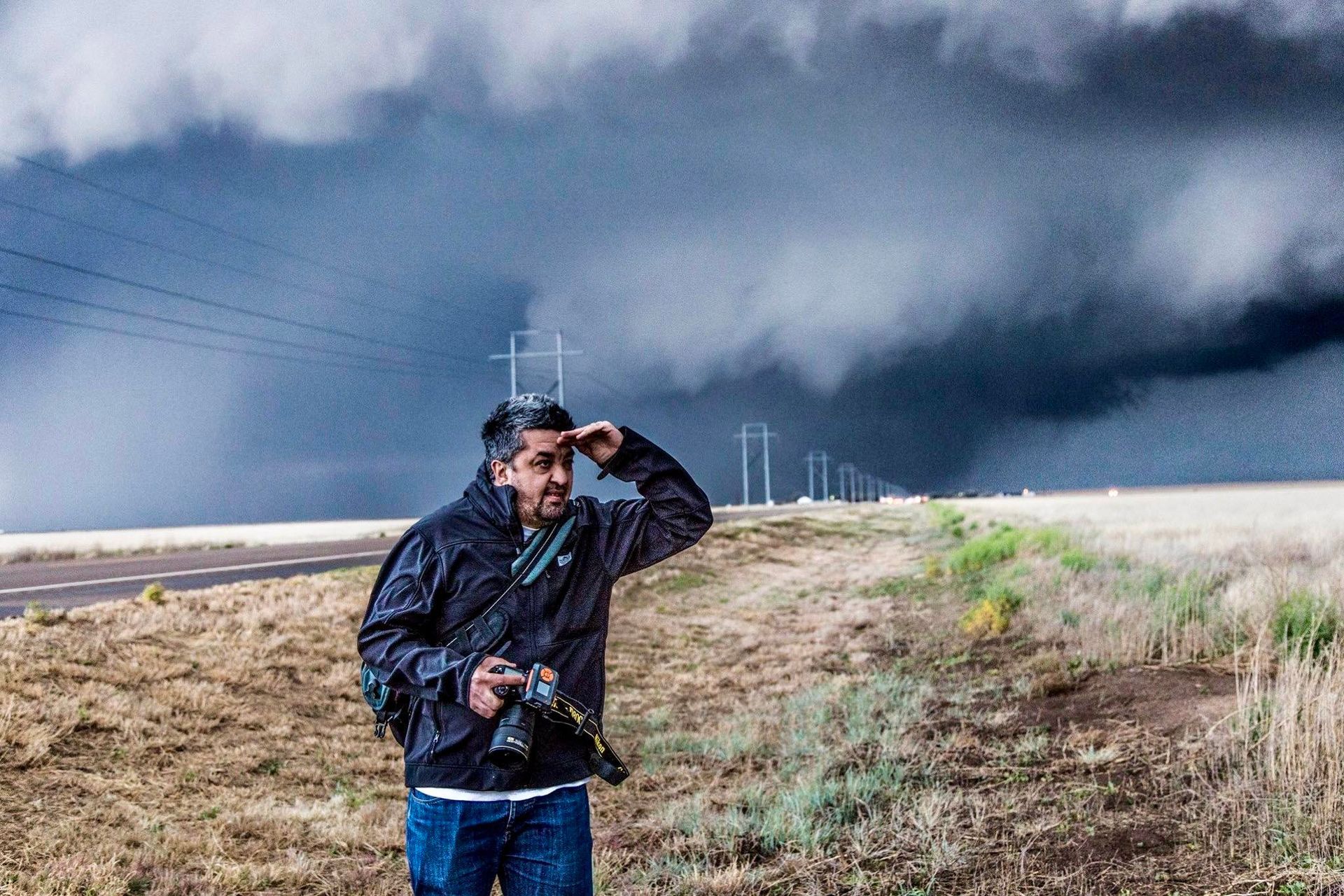 A man is standing in a field holding a camera in front of a storm.