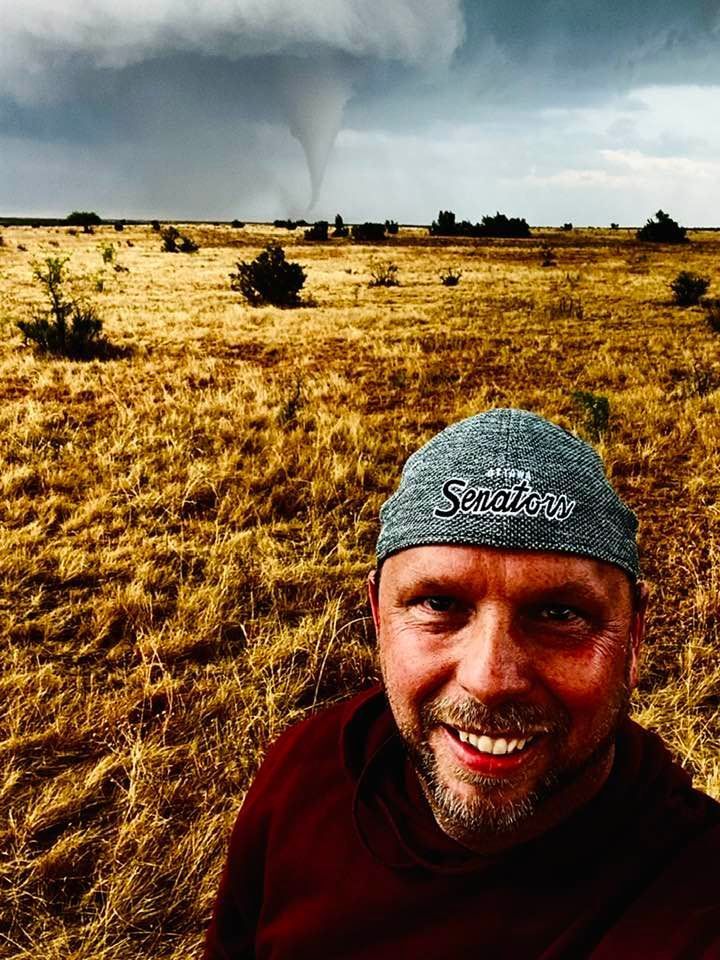 A man is smiling in front of a tornado in a field.
