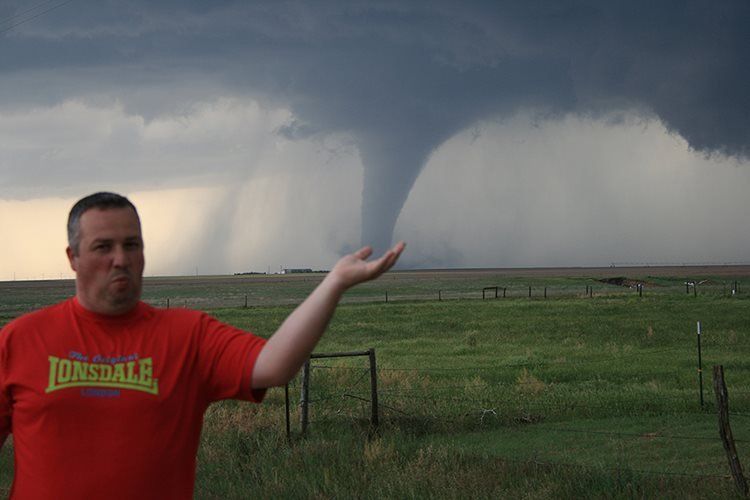 A man in a red lonsdale shirt is standing in front of a tornado.