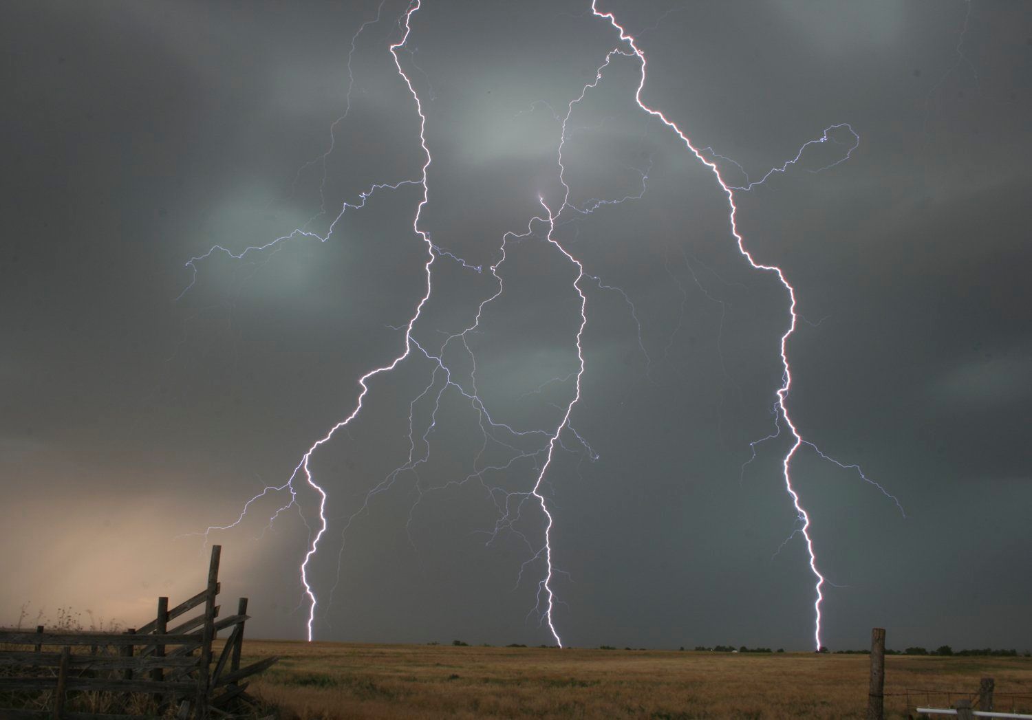 Lightning strikes over a field with a fence in the foreground