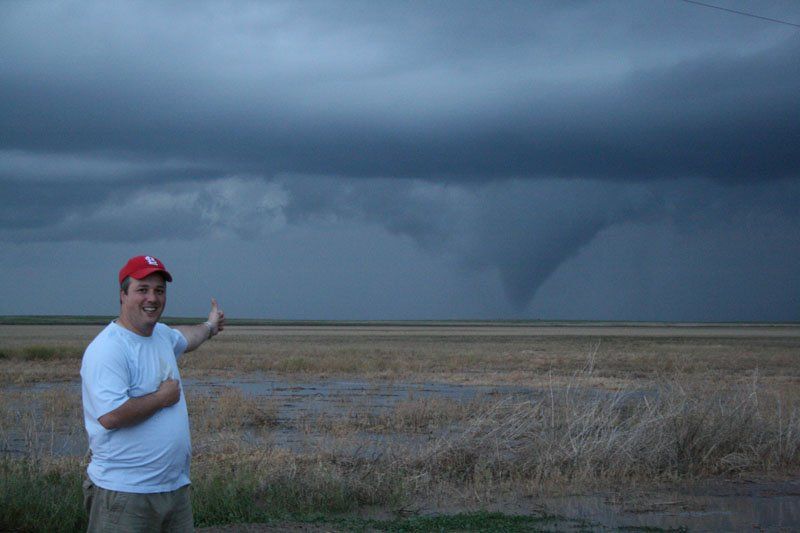 A man is standing in a field pointing at a tornado in the distance.