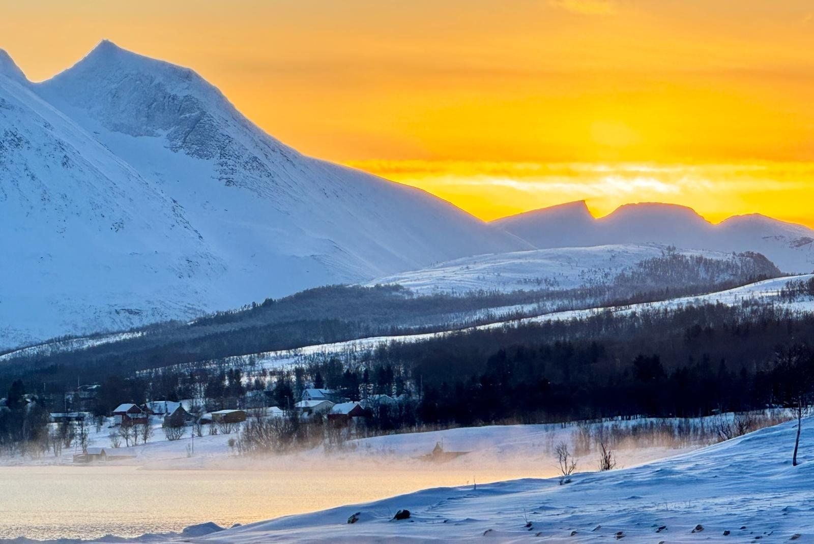 A sunset over a snowy mountain range with a lake in the foreground.