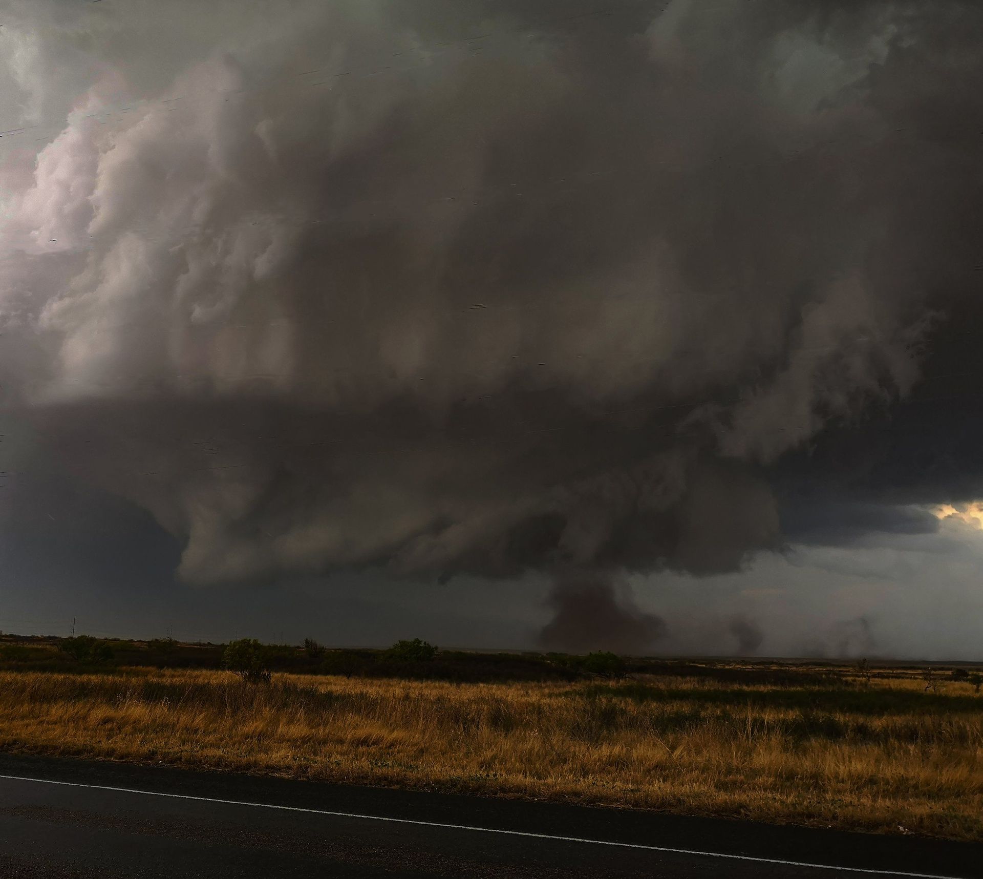 A large storm cloud is moving over a field and a road.