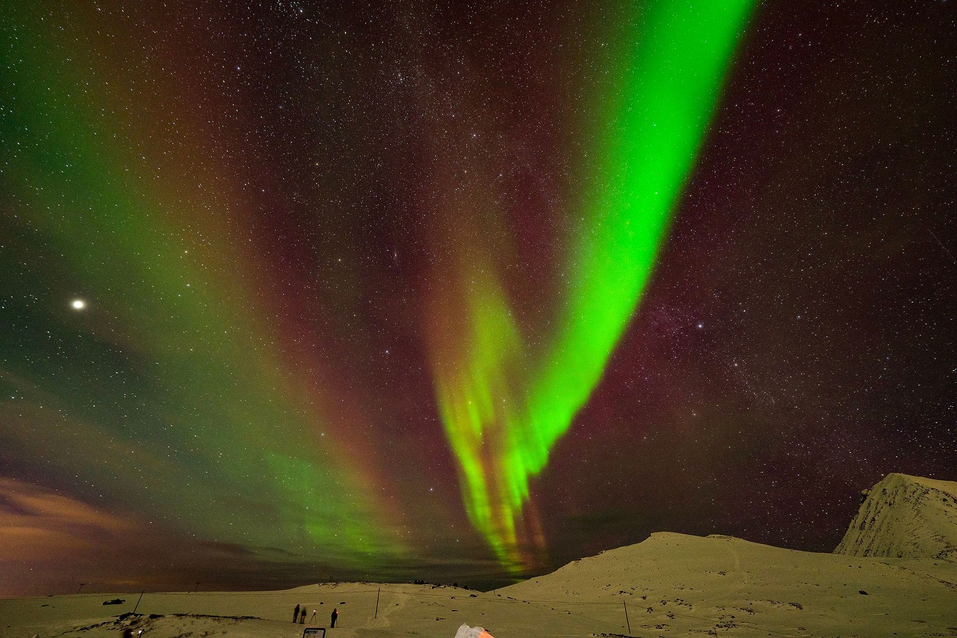 A person is standing in front of a green aurora borealis in the night sky.