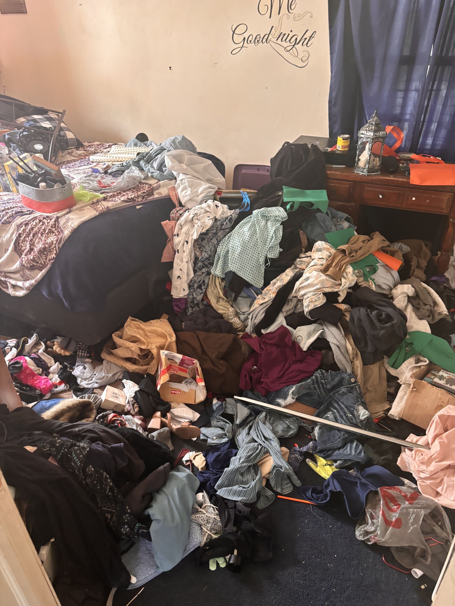 A bedroom with a lot of clothes on the floor and a bed.