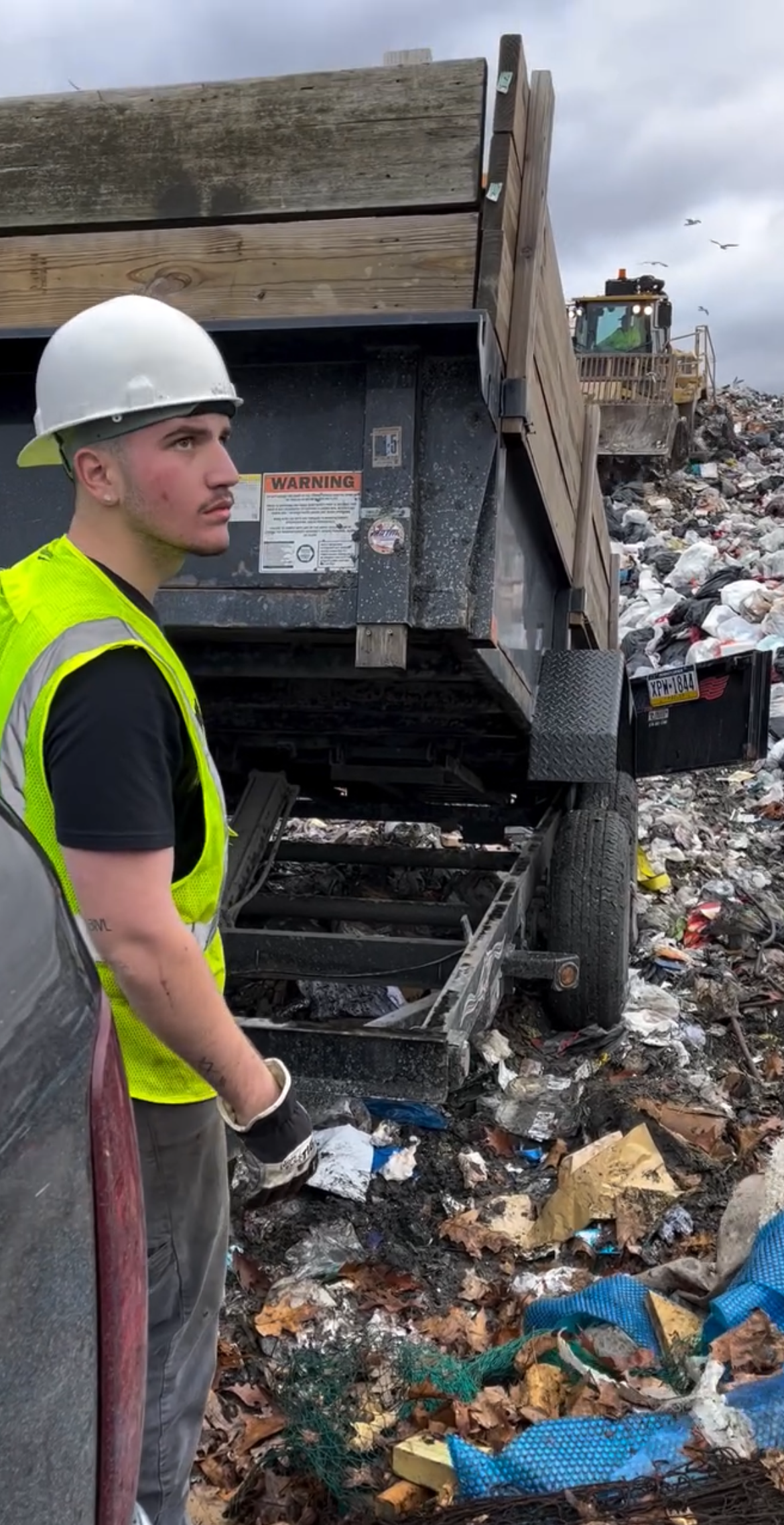 A man wearing a hard hat and safety vest is standing in front of a dumpster.