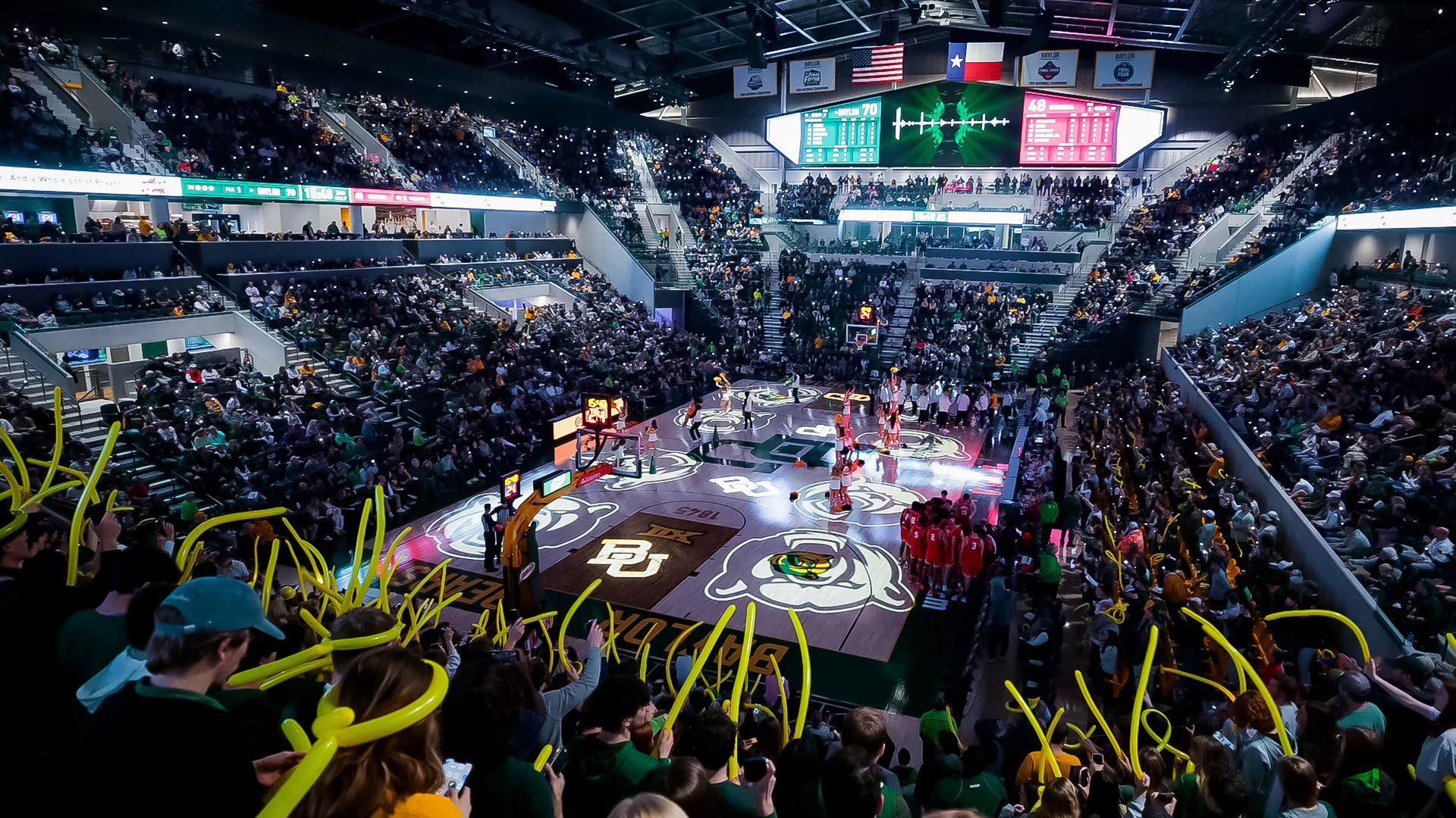 A crowded basketball arena with players on the court and fans cheering, holding yellow foam sticks.