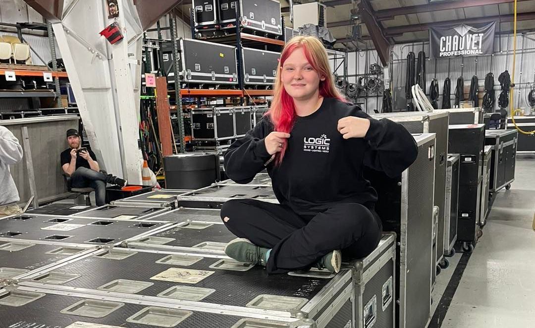 Woman with pink hair sits on cases, smiles in a warehouse setting.