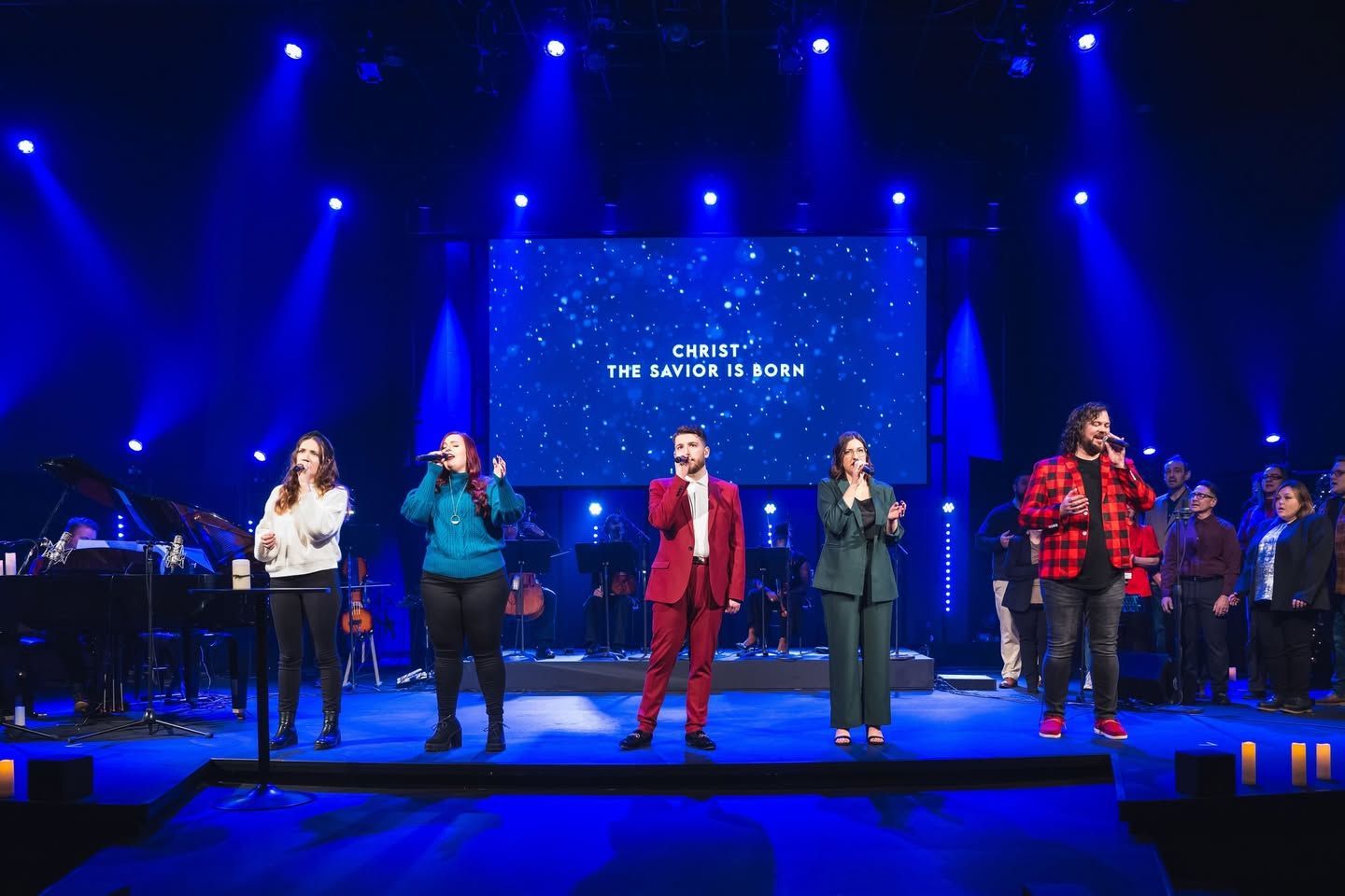 Choir singing on stage, bathed in blue light. Singers in various outfits, screen behind them reads