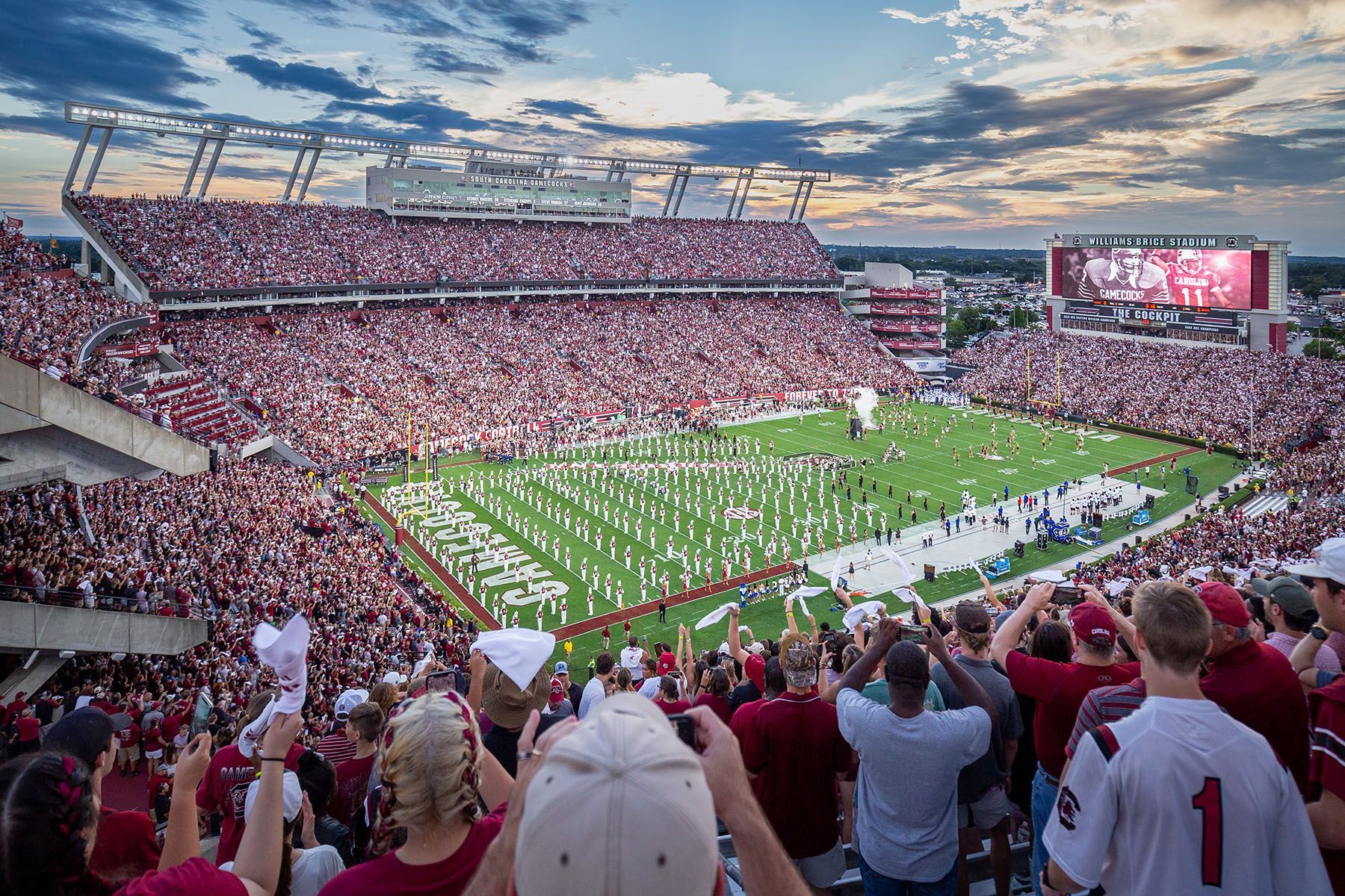 Football stadium packed with fans, field in center, red and white colors prominent, evening sky.