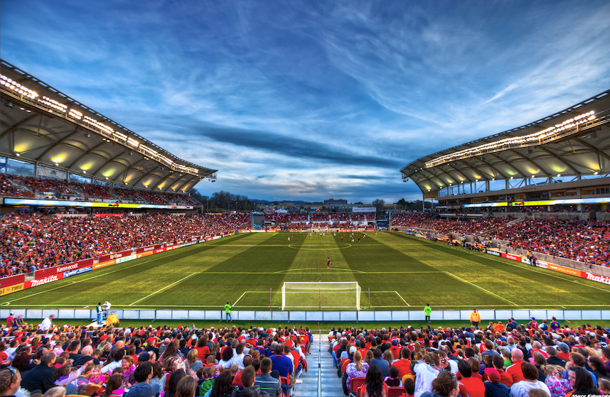 Soccer stadium filled with fans watching a game under a cloudy blue sky.