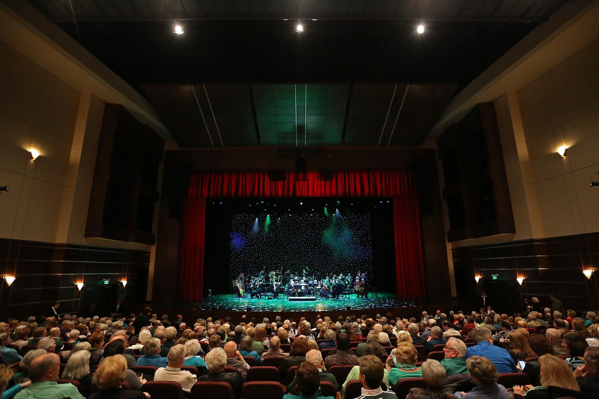 Audience watches a performance on stage at a theater. Red curtains frame the lit stage.