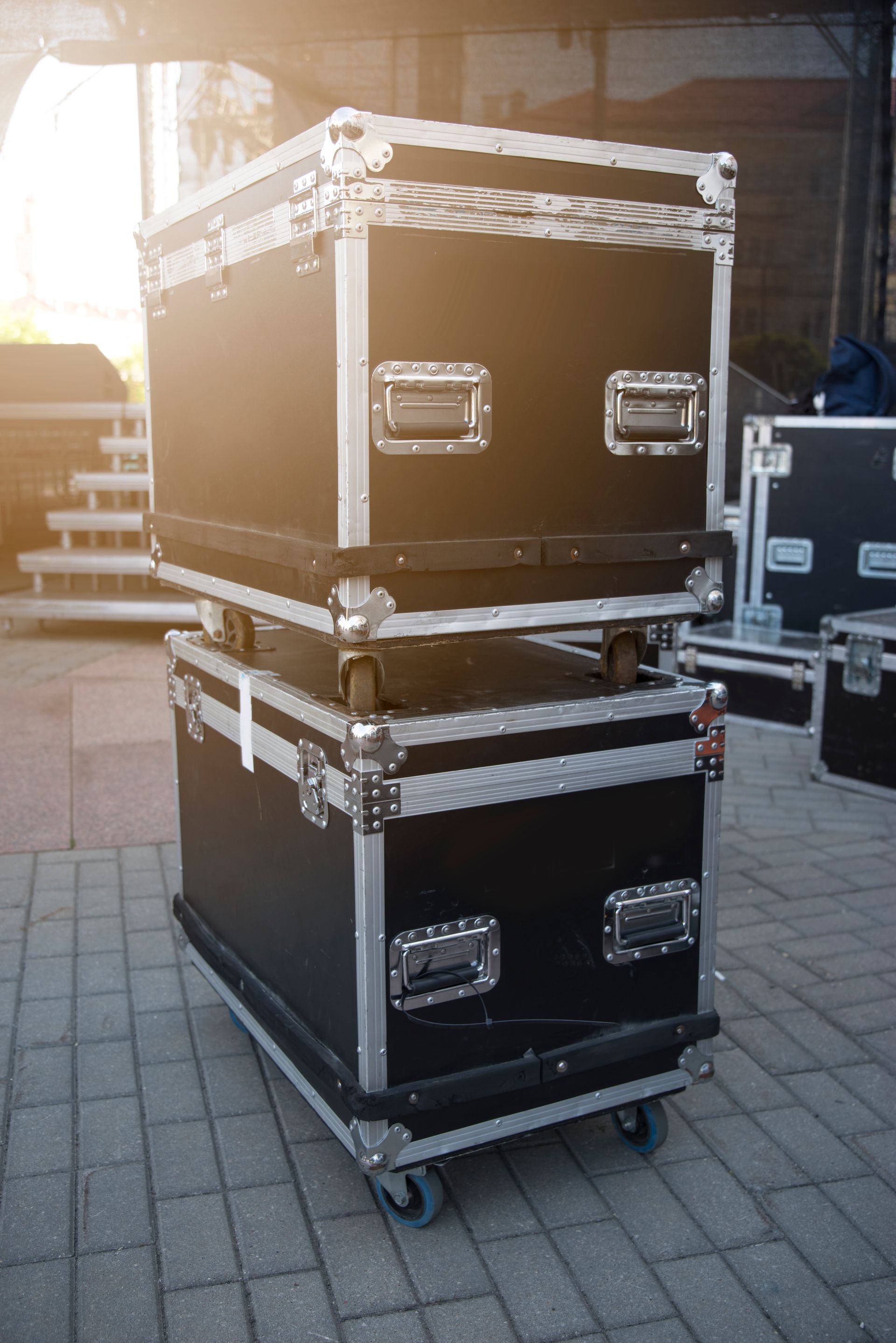 Three black road cases stacked on a brick surface; the bottom one has wheels.