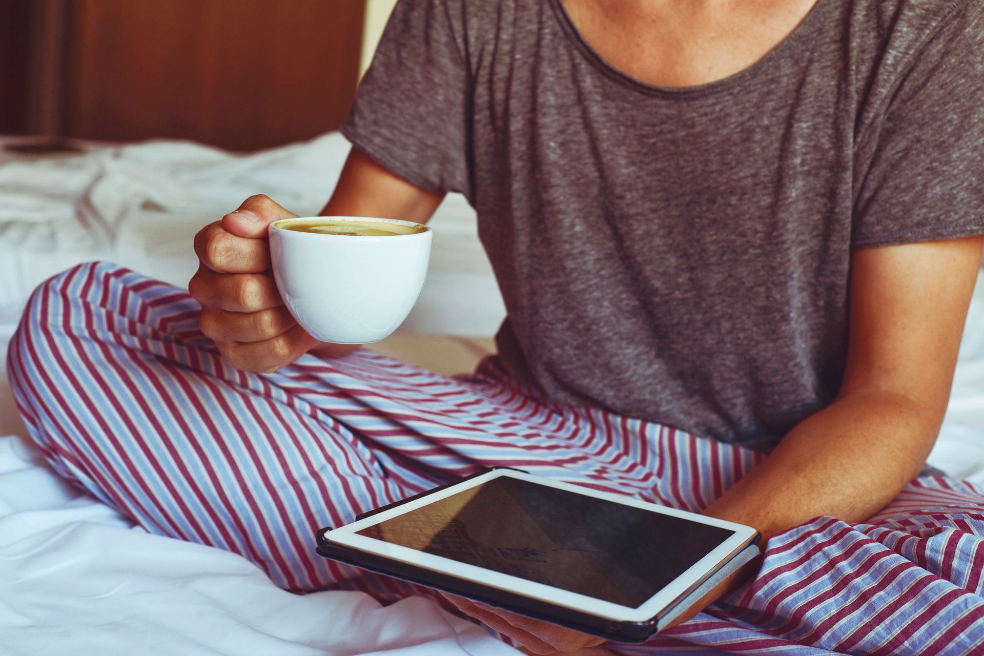 Persona en pijama en la cama, sosteniendo una taza de café y una tableta.
