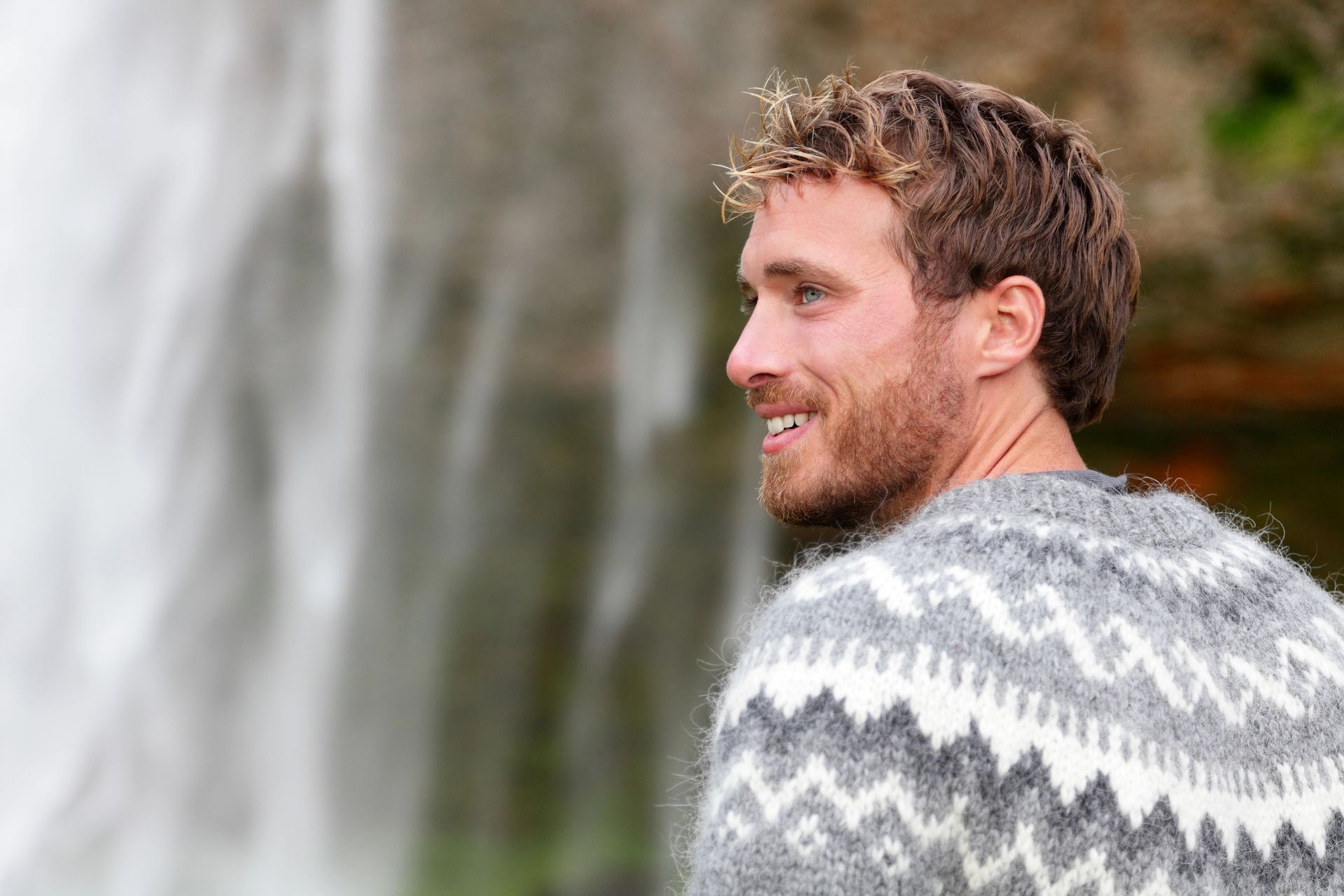 Un hombre con cabello rizado y barba que viste un suéter gris estampado sonríe cerca de una cascada.