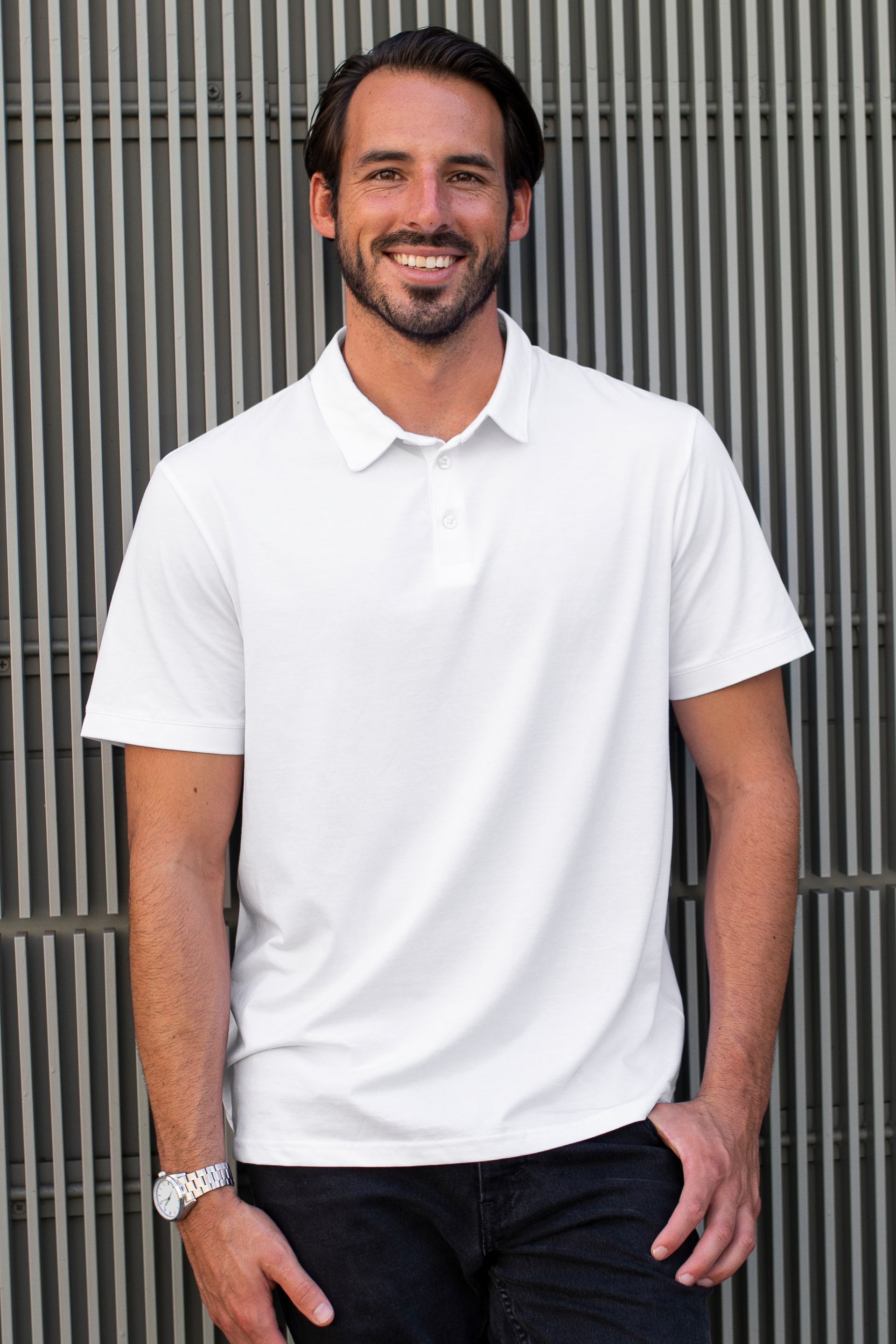 Hombre con polo blanco y jeans oscuros, sonriendo, al aire libre.