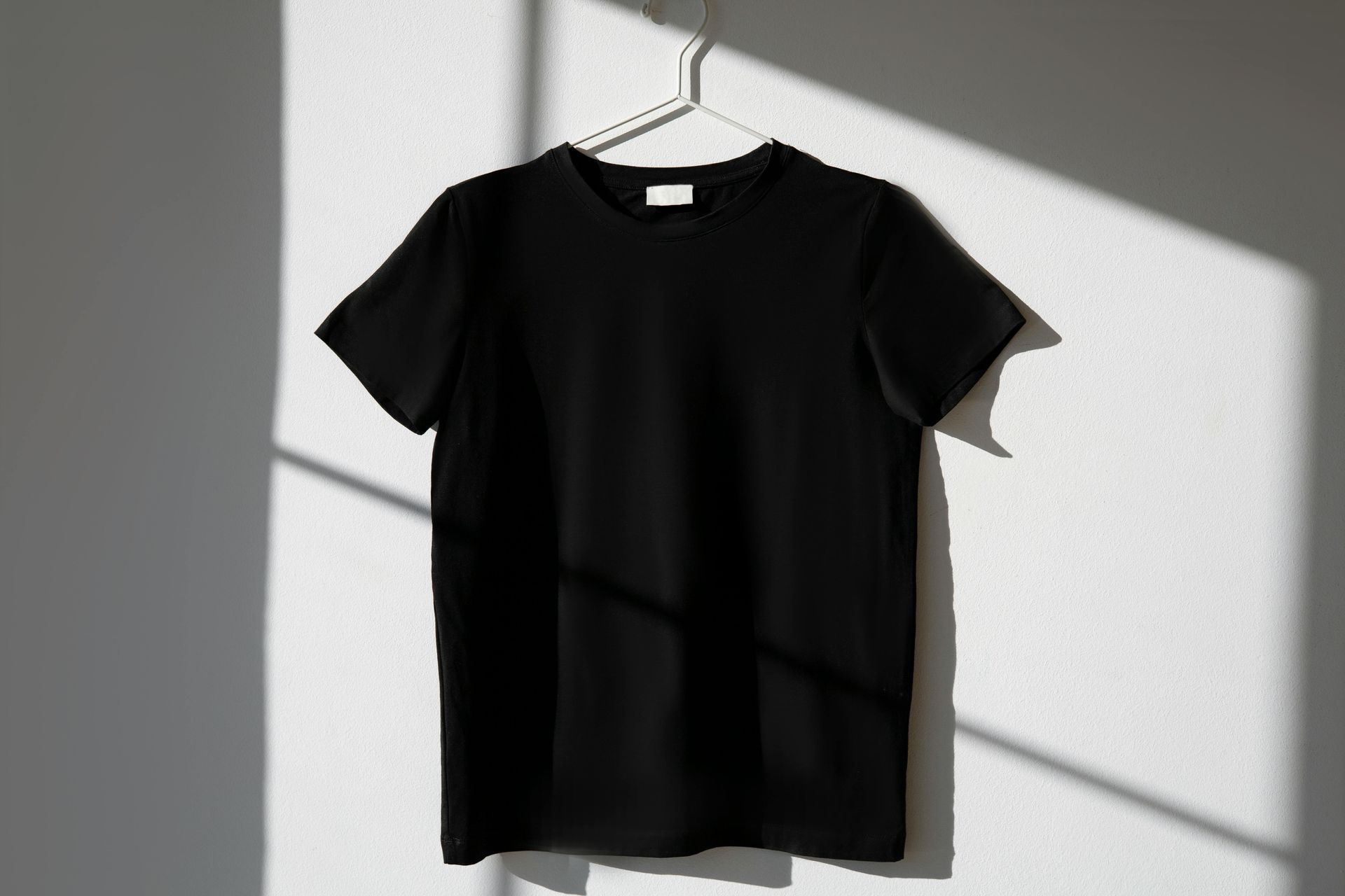 Camiseta negra colgada en una percha blanca contra una pared blanca, con sombra de la luz del sol.