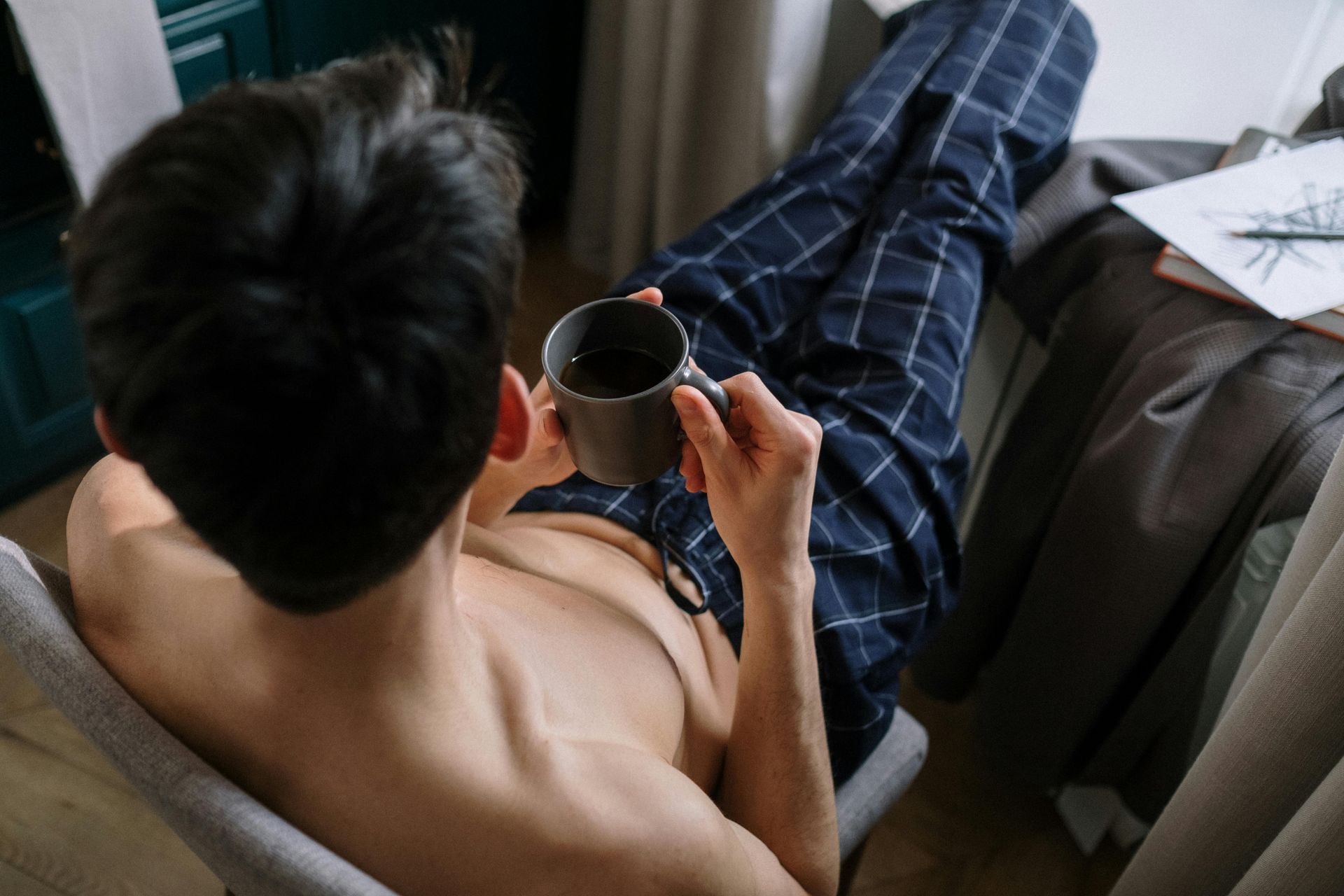 Hombre con pantalones a cuadros, sin camisa, descansando en una silla y sosteniendo una taza de café.