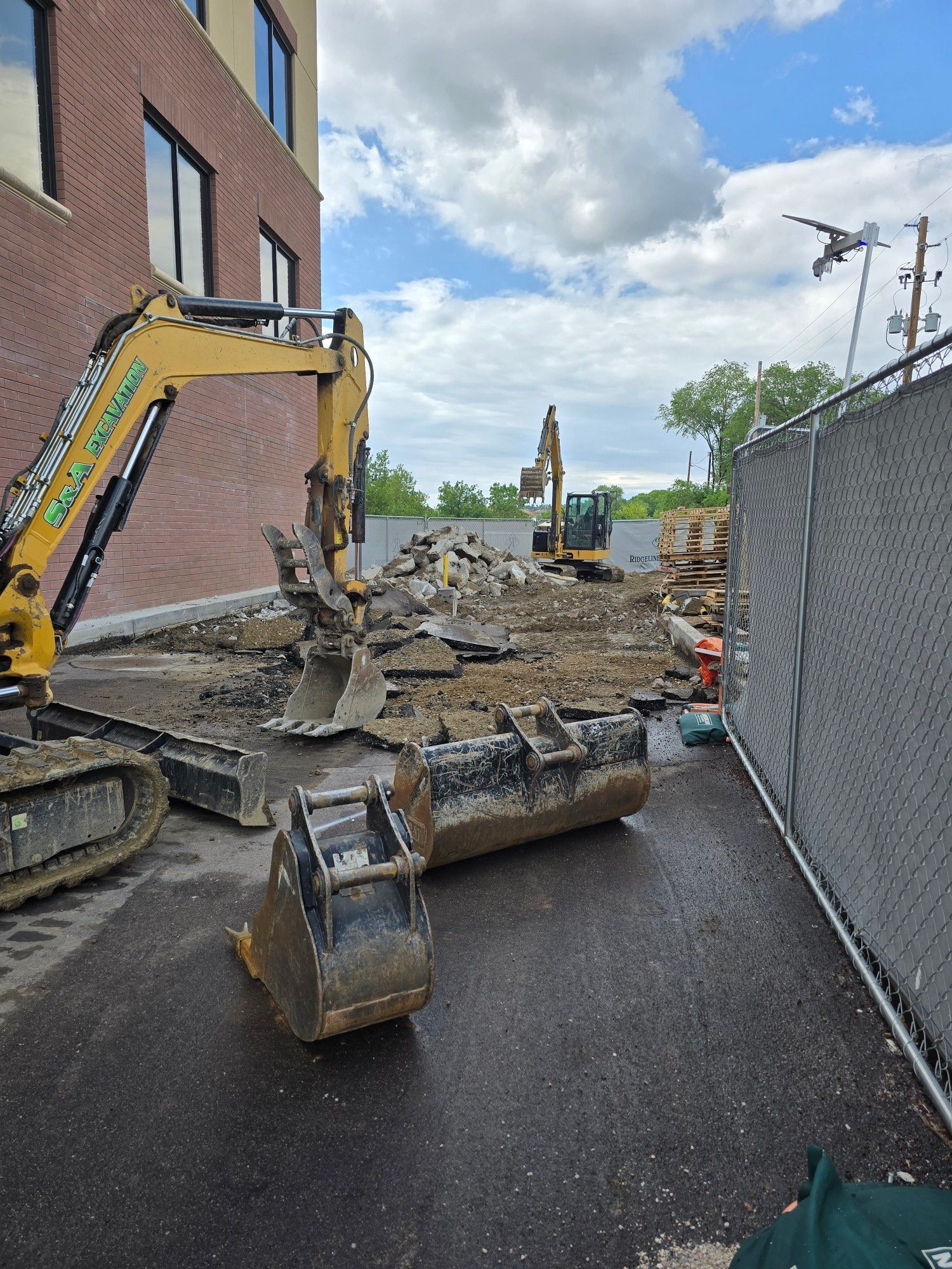 Yellow excavator on a narrow paved path beside a brick building and chain-link fence at a construction site