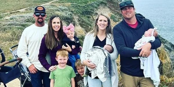 Family of six outdoors on a cloudy trail, smiling and holding two infants, with two dogs in the foreground