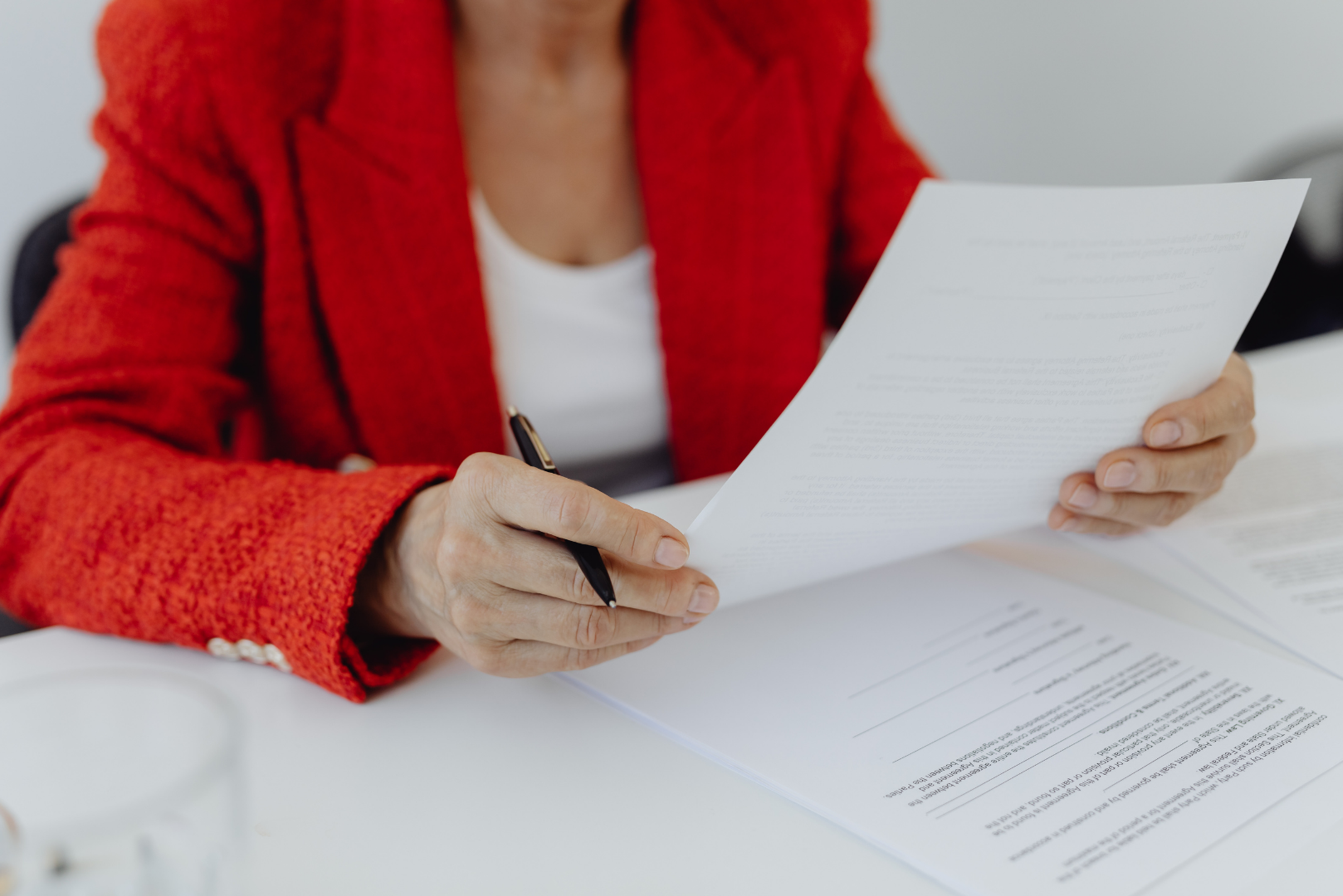 Woman in red blazer holding and reviewing papers, possibly documents, at a white desk.