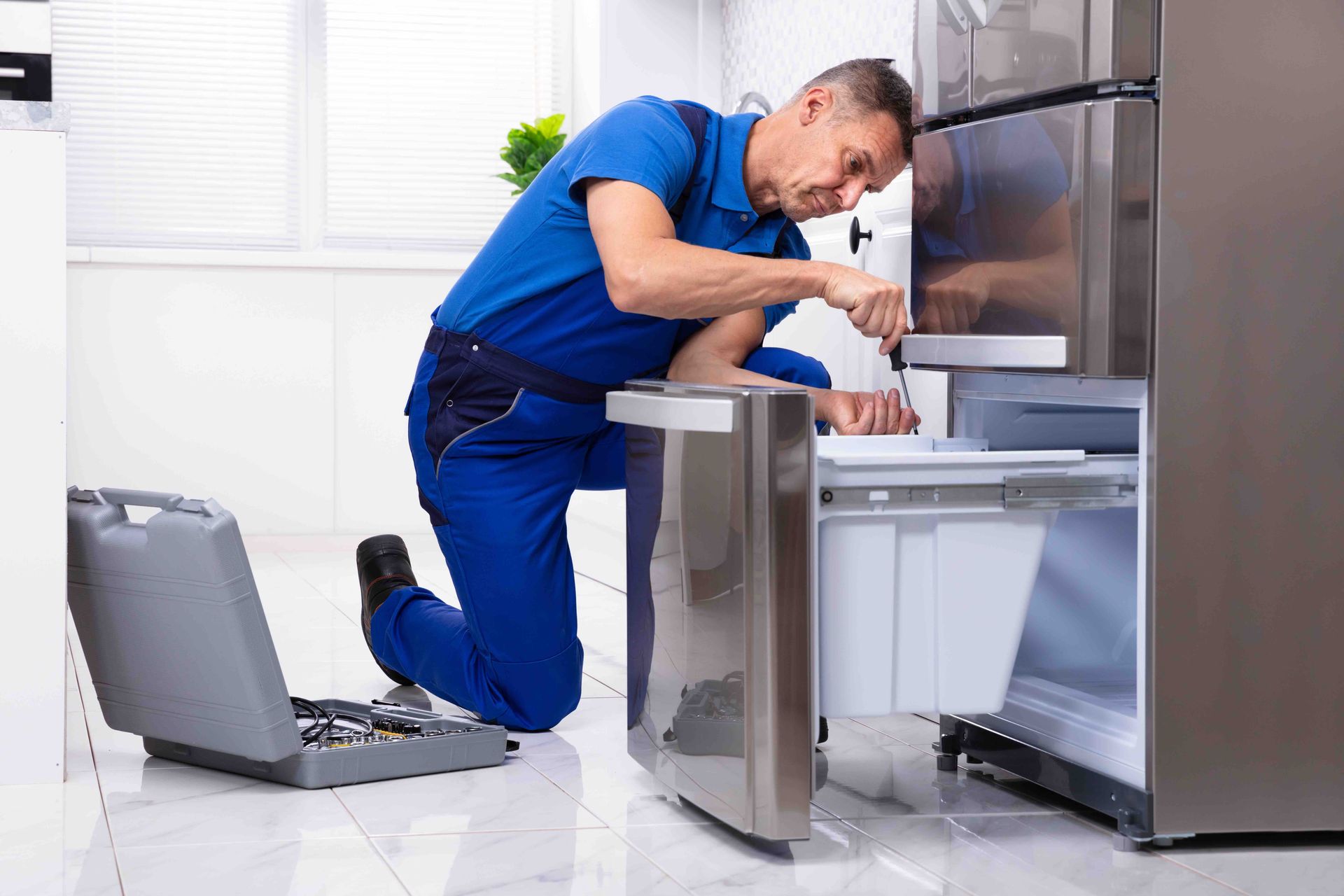 Technician in blue uniform repairing an open stainless steel appliance in a kitchen with tools nearby