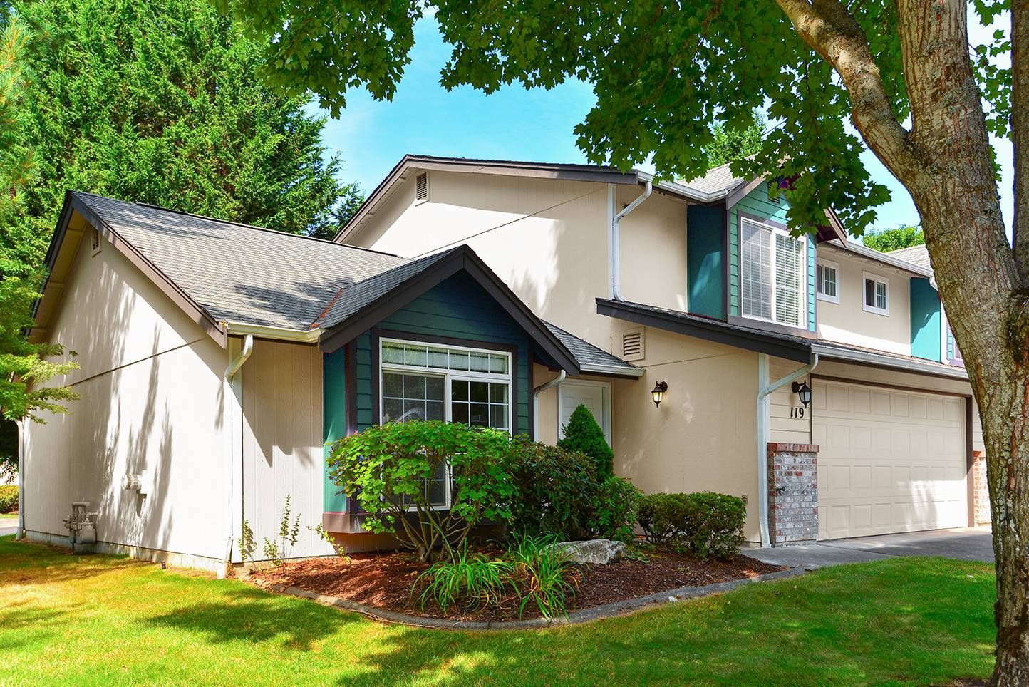 Exterior view of a beige townhouse with teal trim, front yard landscaping, and a two-car garage.