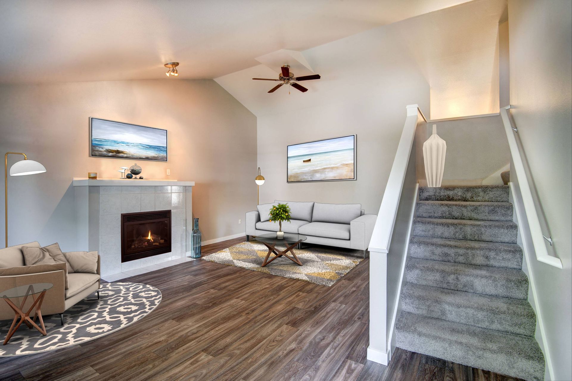 Interior living room with gray sofa, fireplace, staircase, ceiling fan, and wood-look flooring.