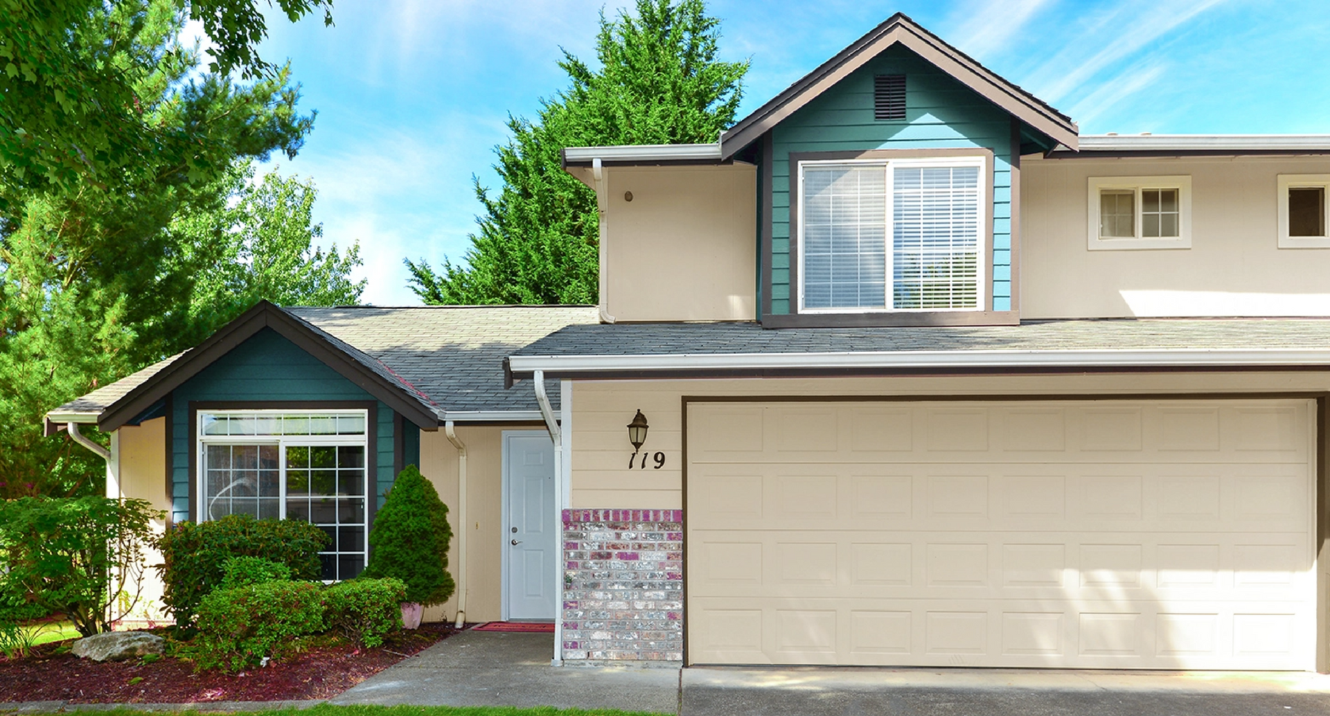 Beige house with teal trim, large garage door, and green lawn on a sunny day.
