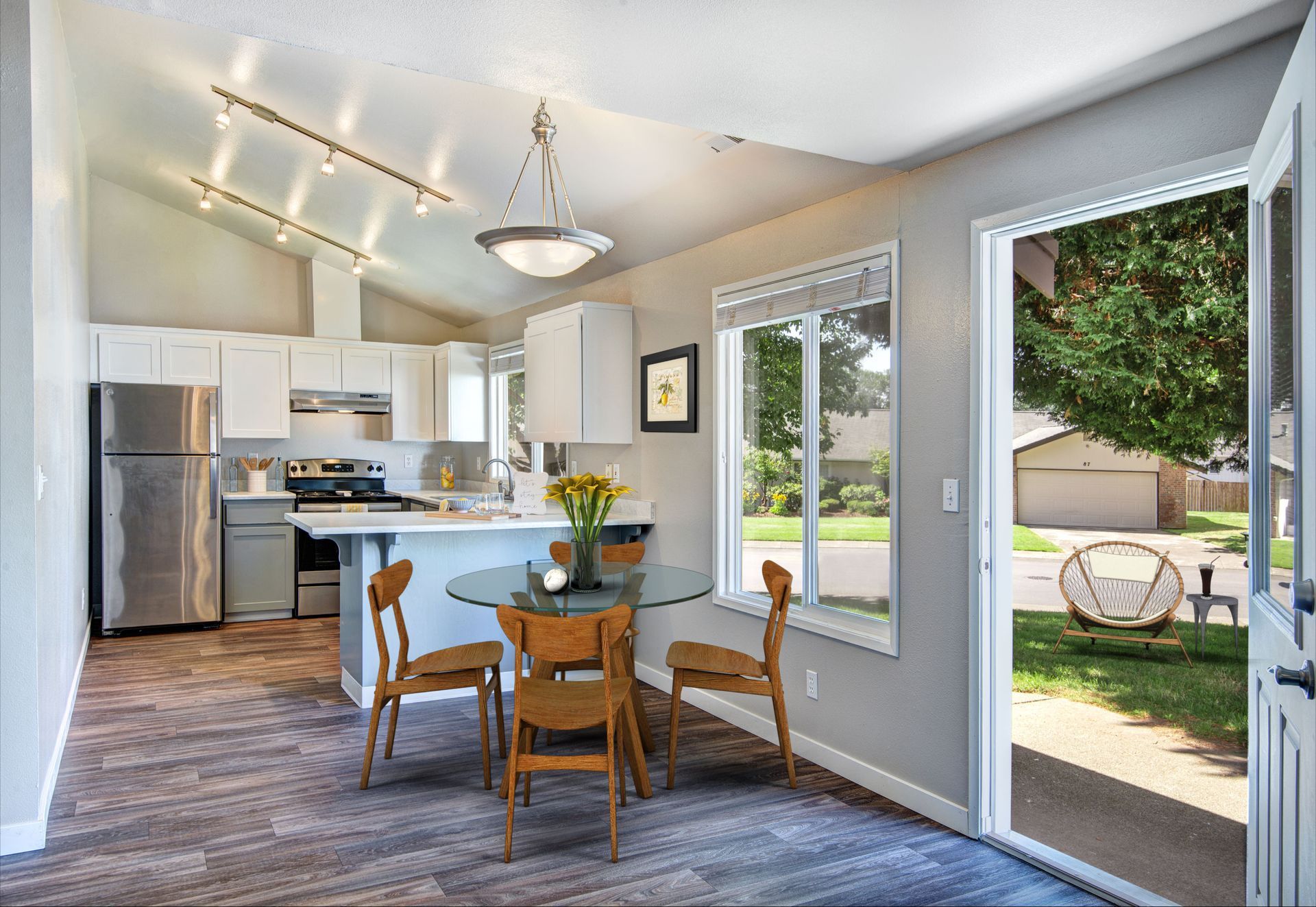 Dining area with round table, chairs, and open doors to a yard. Kitchen in background, stainless steel appliances, neutral tones.