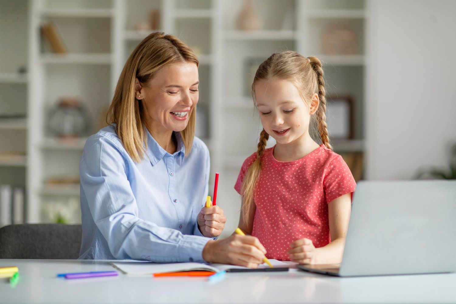 Woman comforting a girl at a desk, near a window and plant, while the girl is writing.