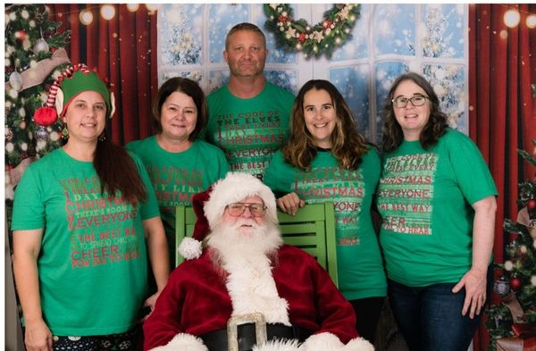 Santa seated with five people in green holiday shirts in front of a festive Christmas backdrop